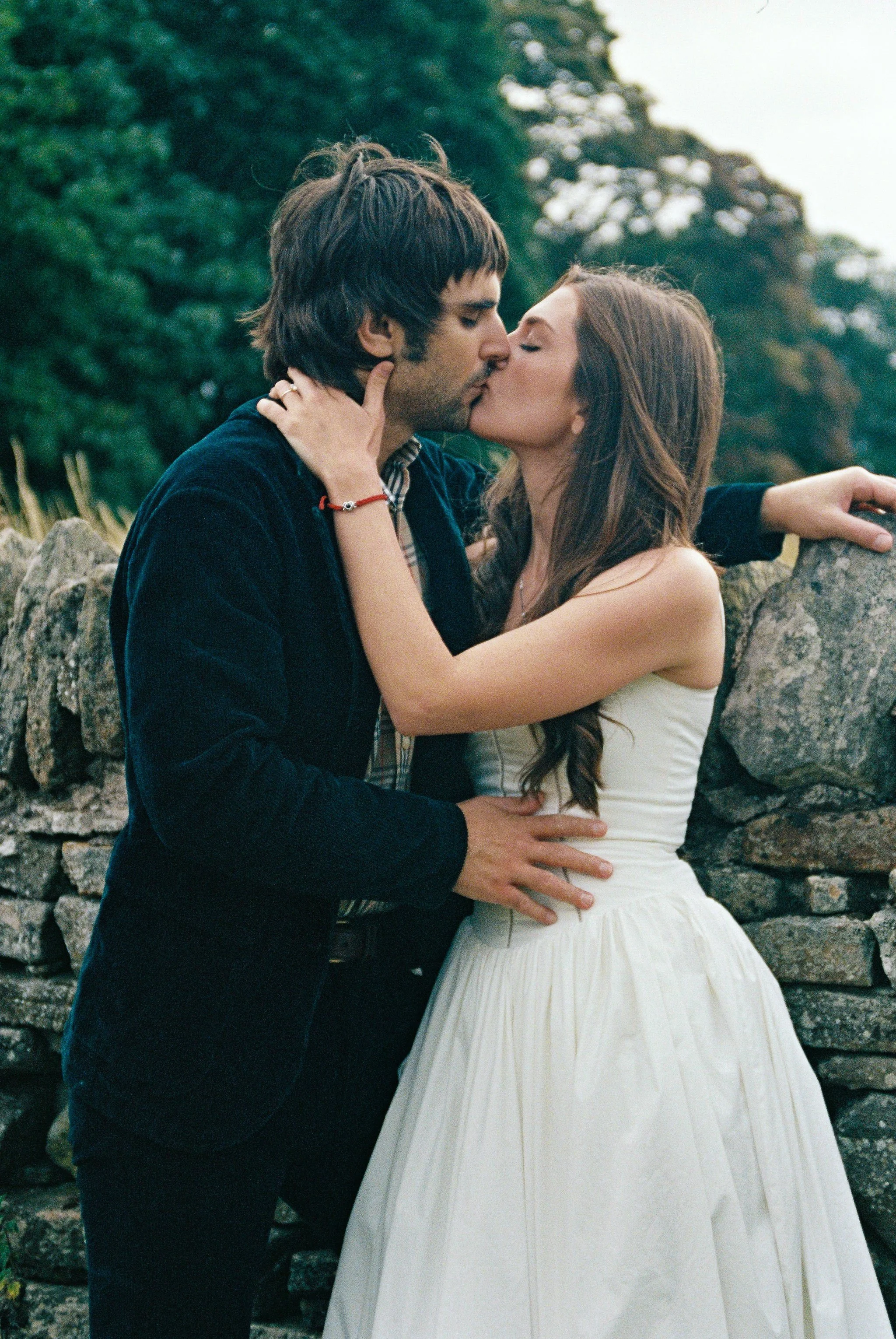 A couple sharing a kiss outdoors next to a stone wall, with lush green trees in the background.