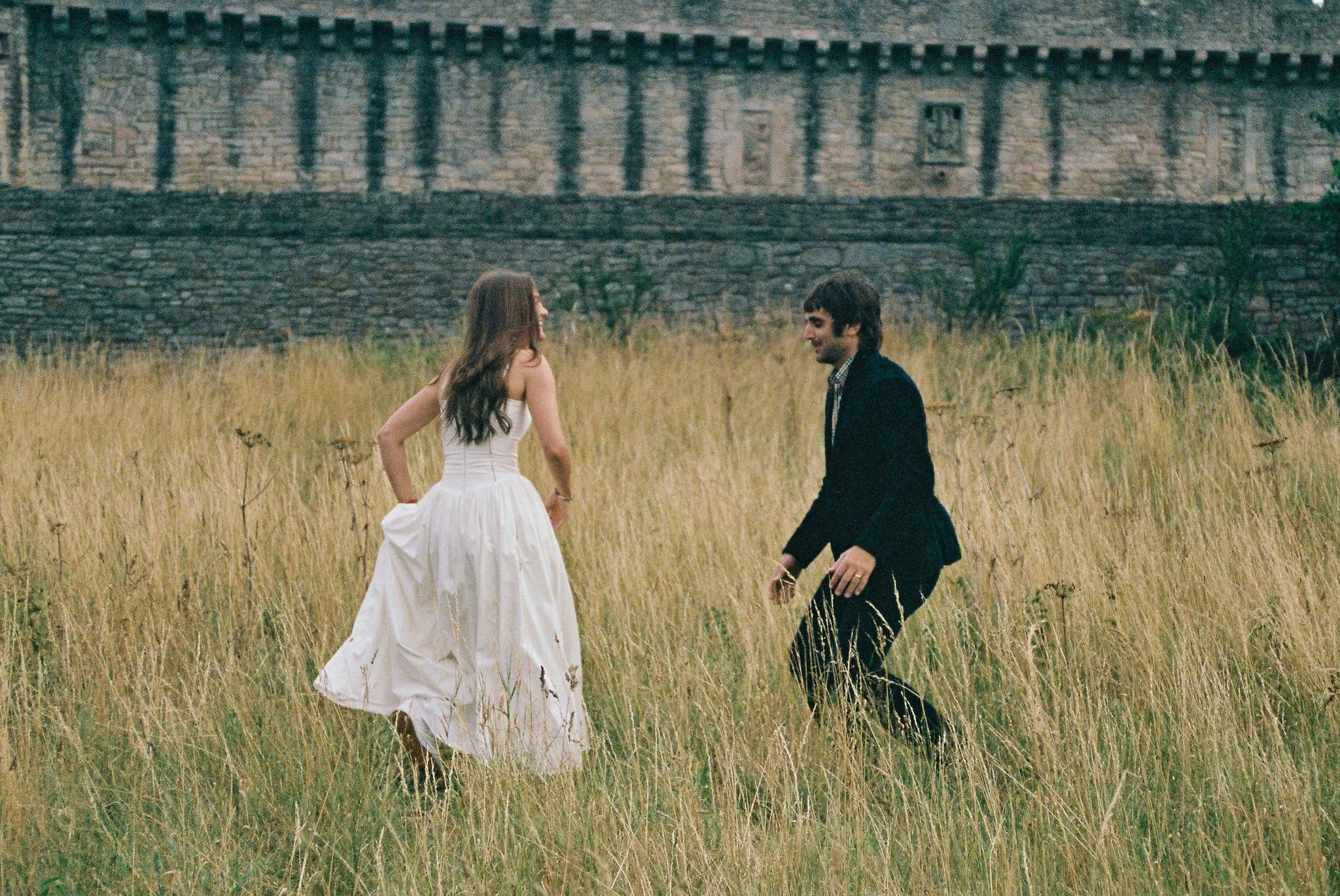 A man and a woman in formal attire standing and dancing in a field of tall grass with an old stone wall in the background.