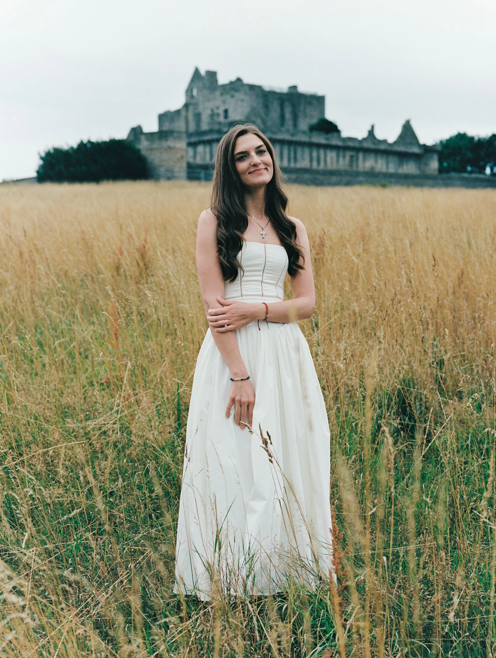 A young woman in a white dress standing in a field of tall grass, with a castle in the background on a cloudy day.