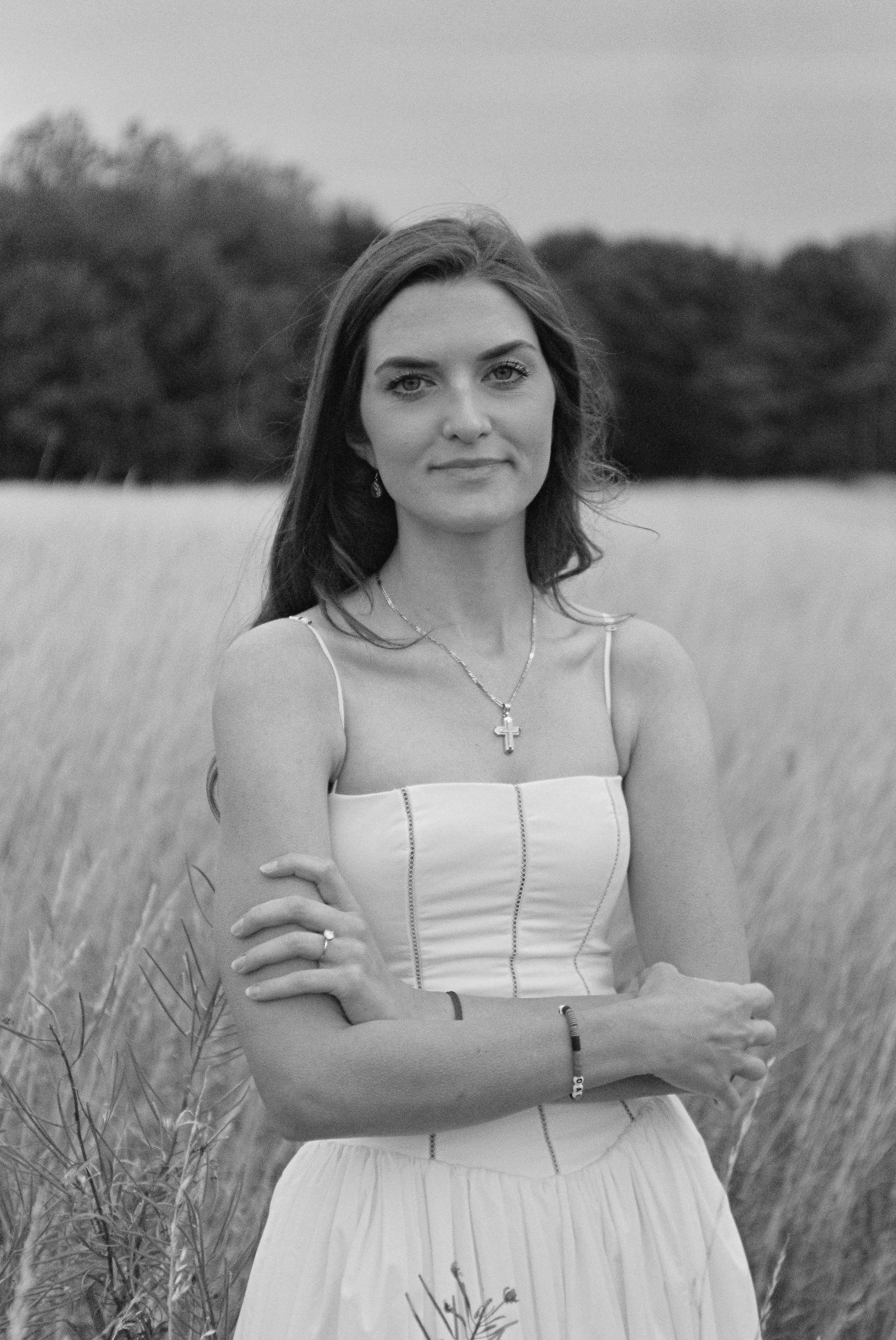 Black and white portrait of a young woman standing in a field, facing the camera with her arms crossed, wearing a sleeveless dress, a cross necklace, and earrings. Background shows trees and tall grass.