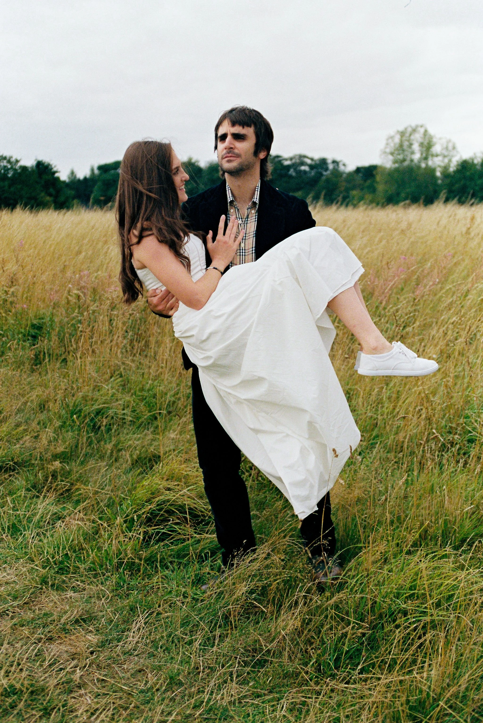 A man wearing dark clothing carries a woman in a white dress and sneakers through a grassy field with trees in the background.