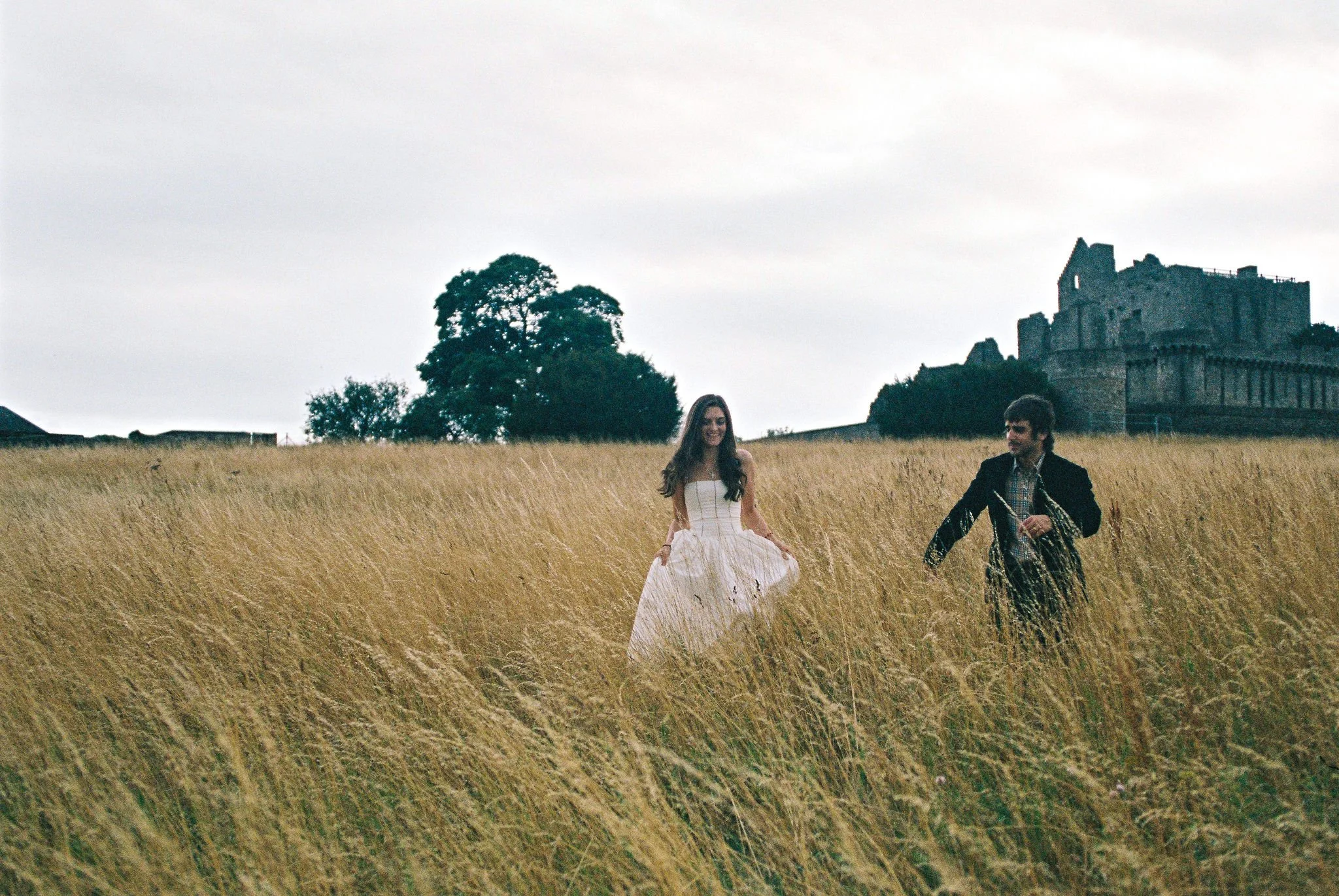 A woman in a white dress and a man in a black jacket walking through a field of tall golden grass near a castle on a cloudy day.