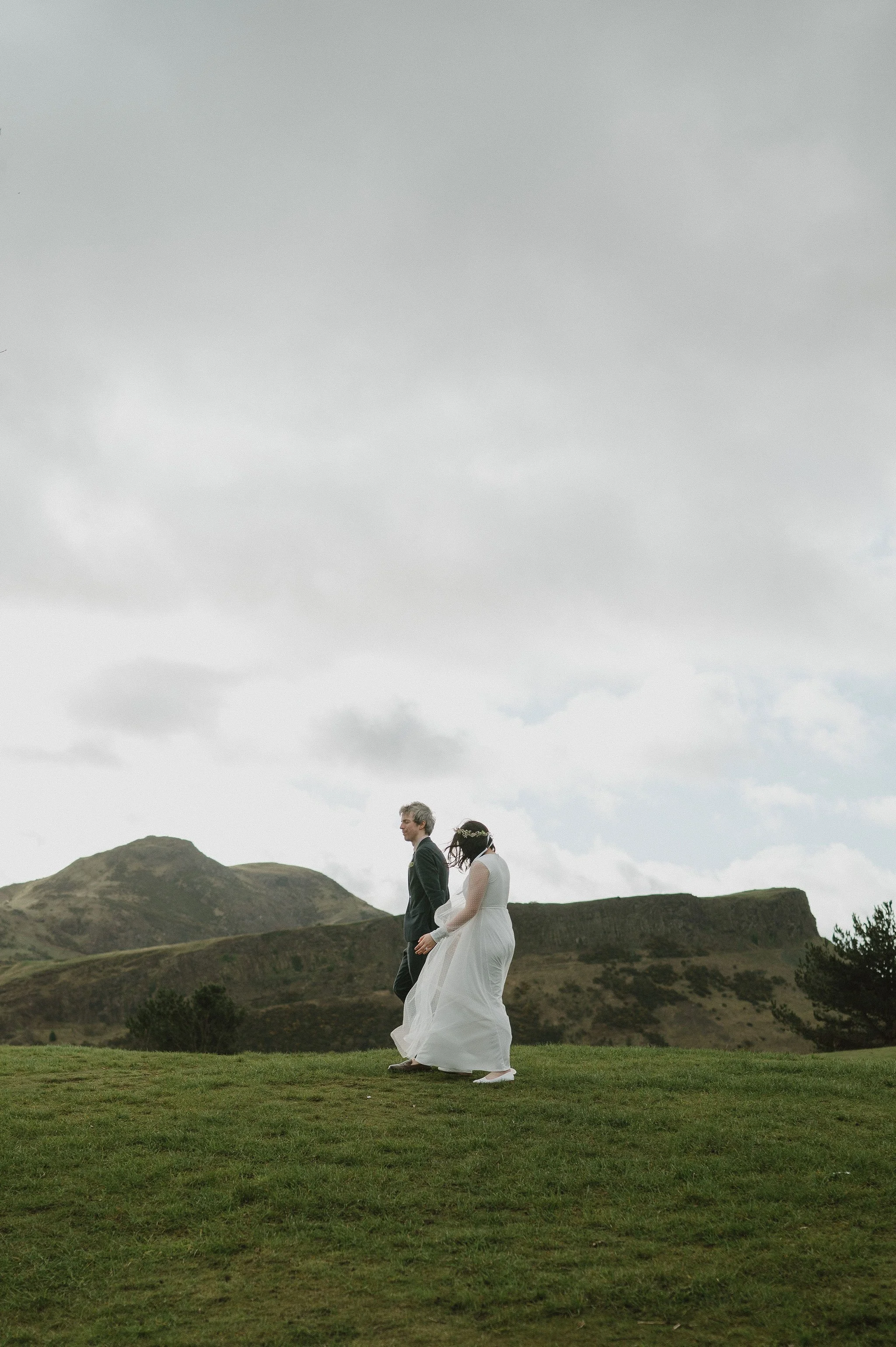 A bride and groom walking hand in hand on a grassy hill with mountains in the background under a cloudy sky.