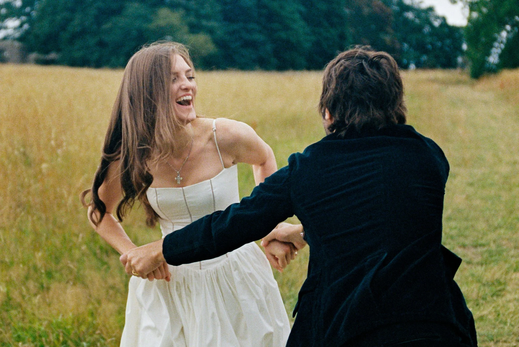 A woman in a white dress laughing and holding hands with a man in a dark jacket in a field.