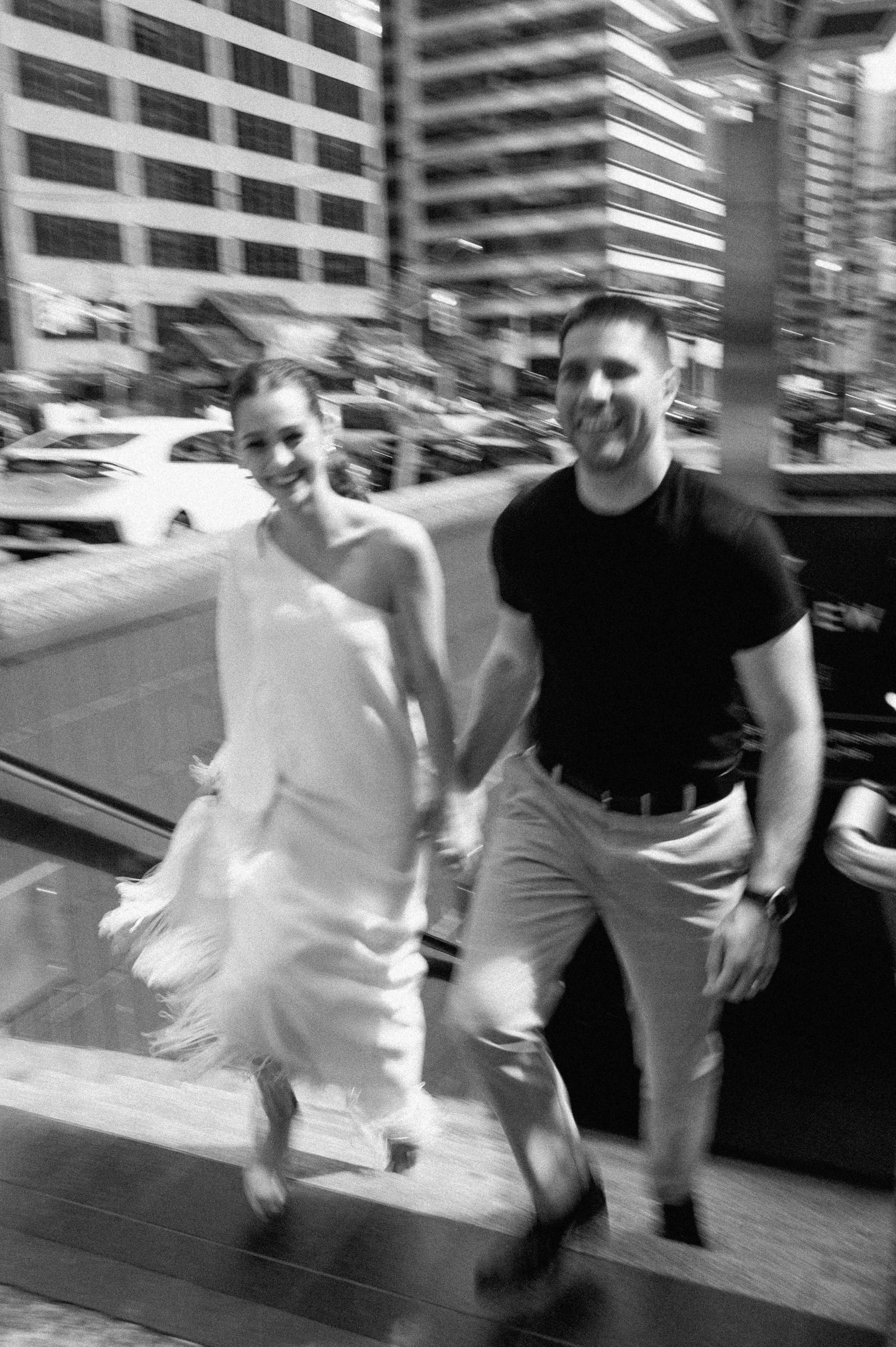 A smiling woman in a white dress holding hands with a smiling man in a black shirt and khaki pants, walking outside on a city street at night, with buildings and parked cars in the background.