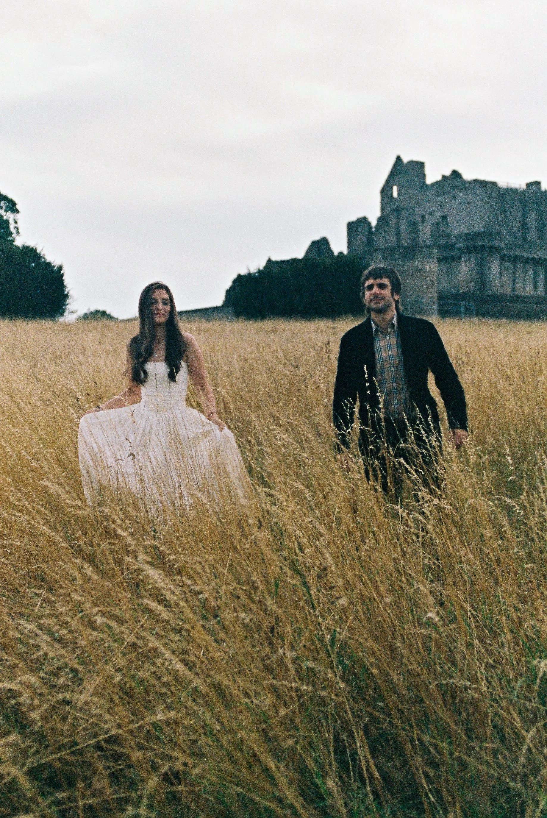 A woman in a white dress and a man in a dark jacket walk through a field of tall grass with the ruins of a castle in the background.