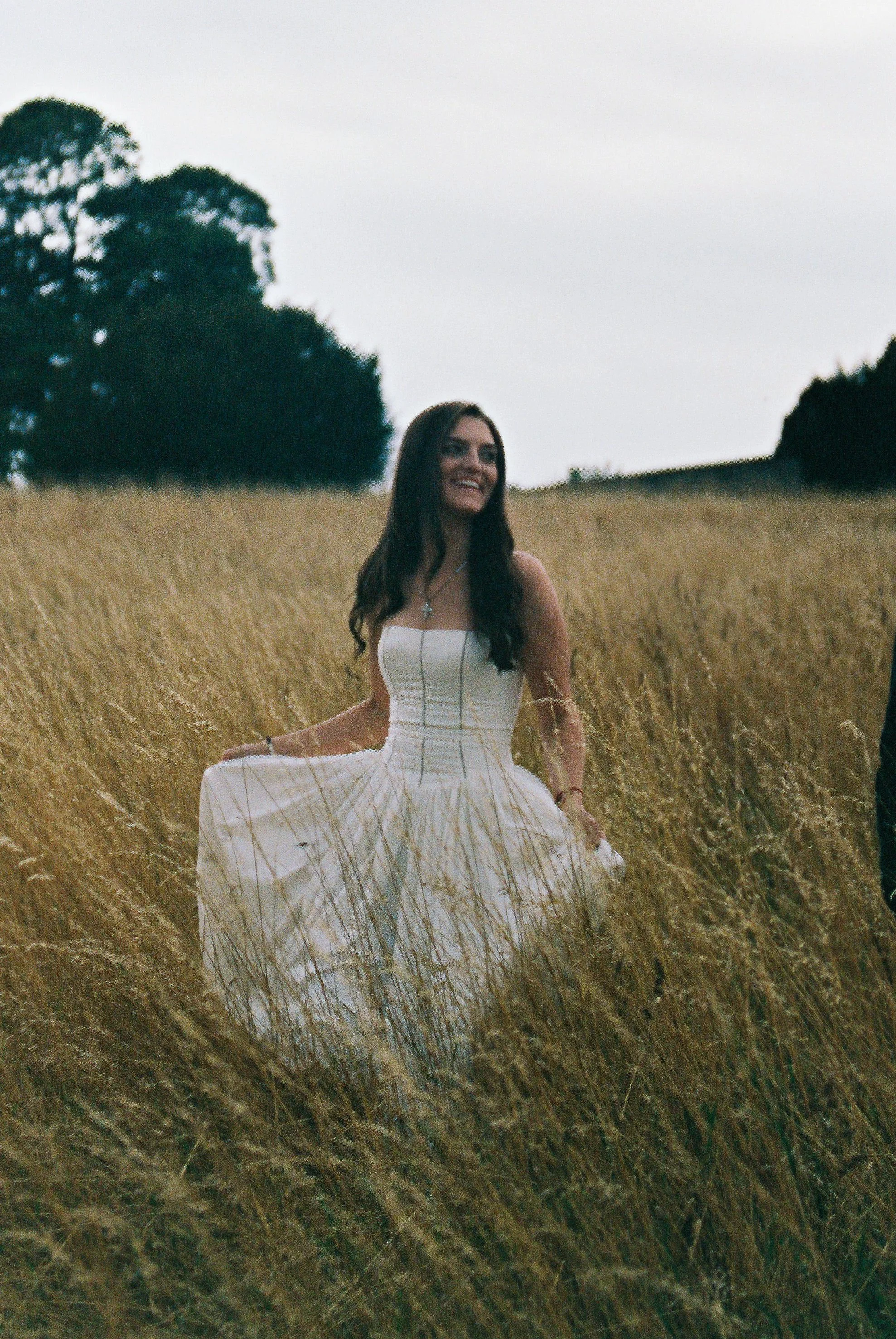 A woman in a white dress standing in a grassy field, smiling, with trees in the background on a cloudy day.