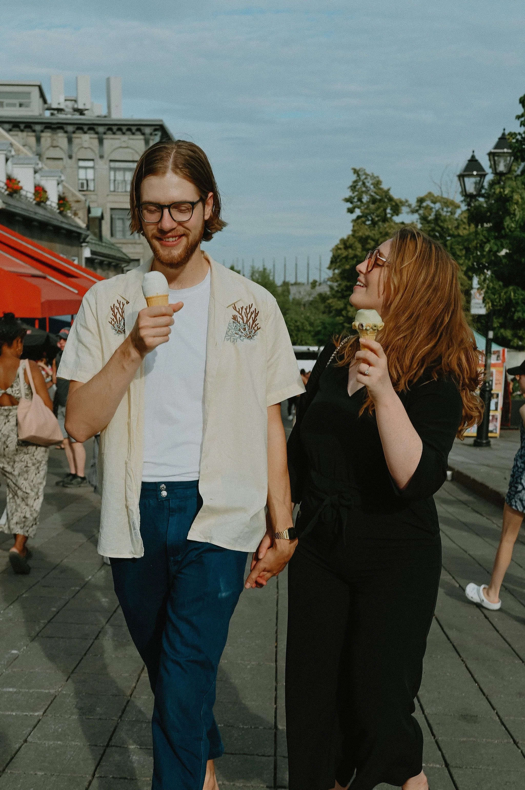 A young couple holding hands and eating ice cream cones while walking outdoors in a city street.