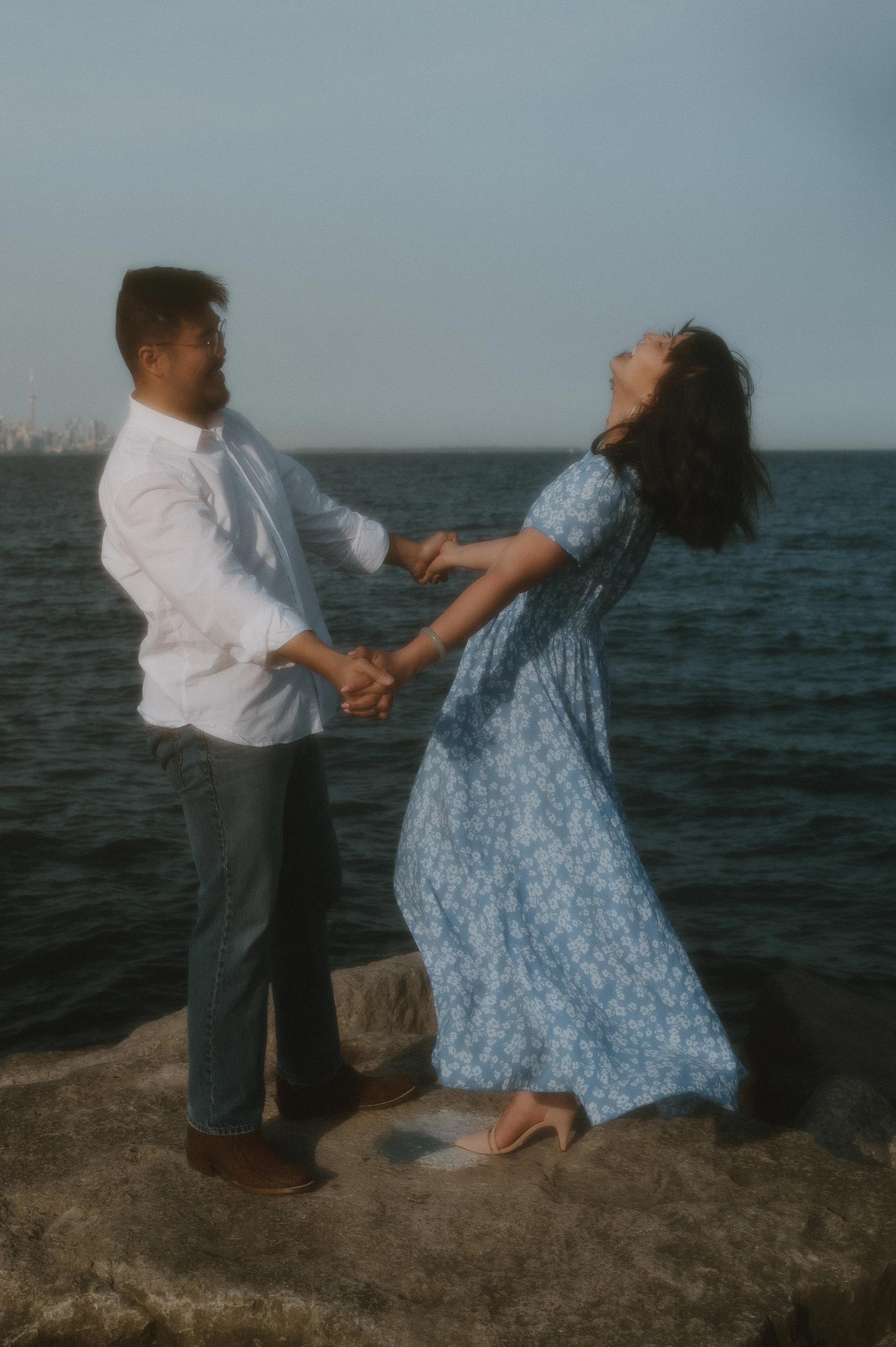 A couple holding hands and smiling on a rocky shore near the water, with the ocean and distant city skyline in the background.
