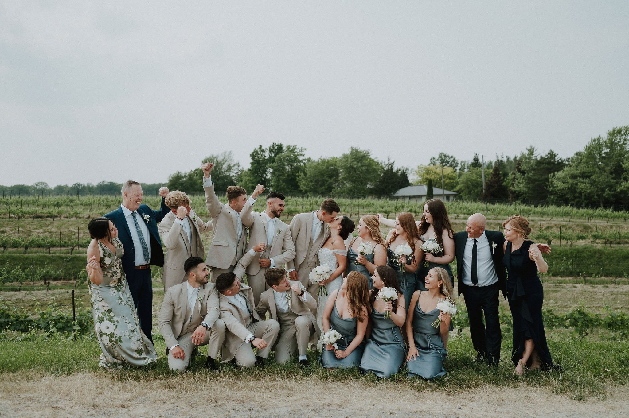 A wedding party outdoors, with the bride and groom kissing, surrounded by friends and family celebrating.