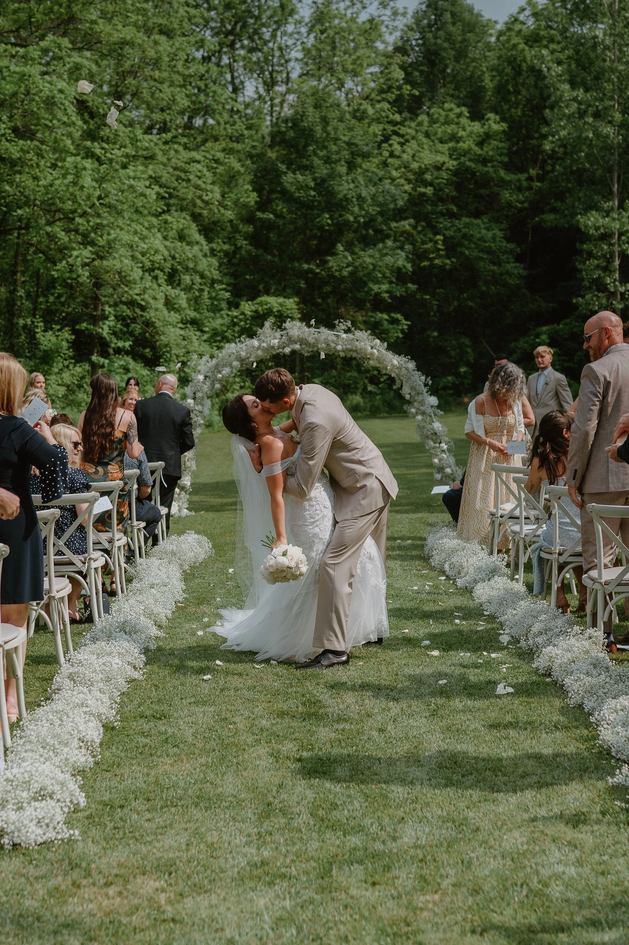 A couple in wedding attire sharing a kiss on an outdoor wedding aisle, surrounded by friends and family, with a floral arch and greenery background.