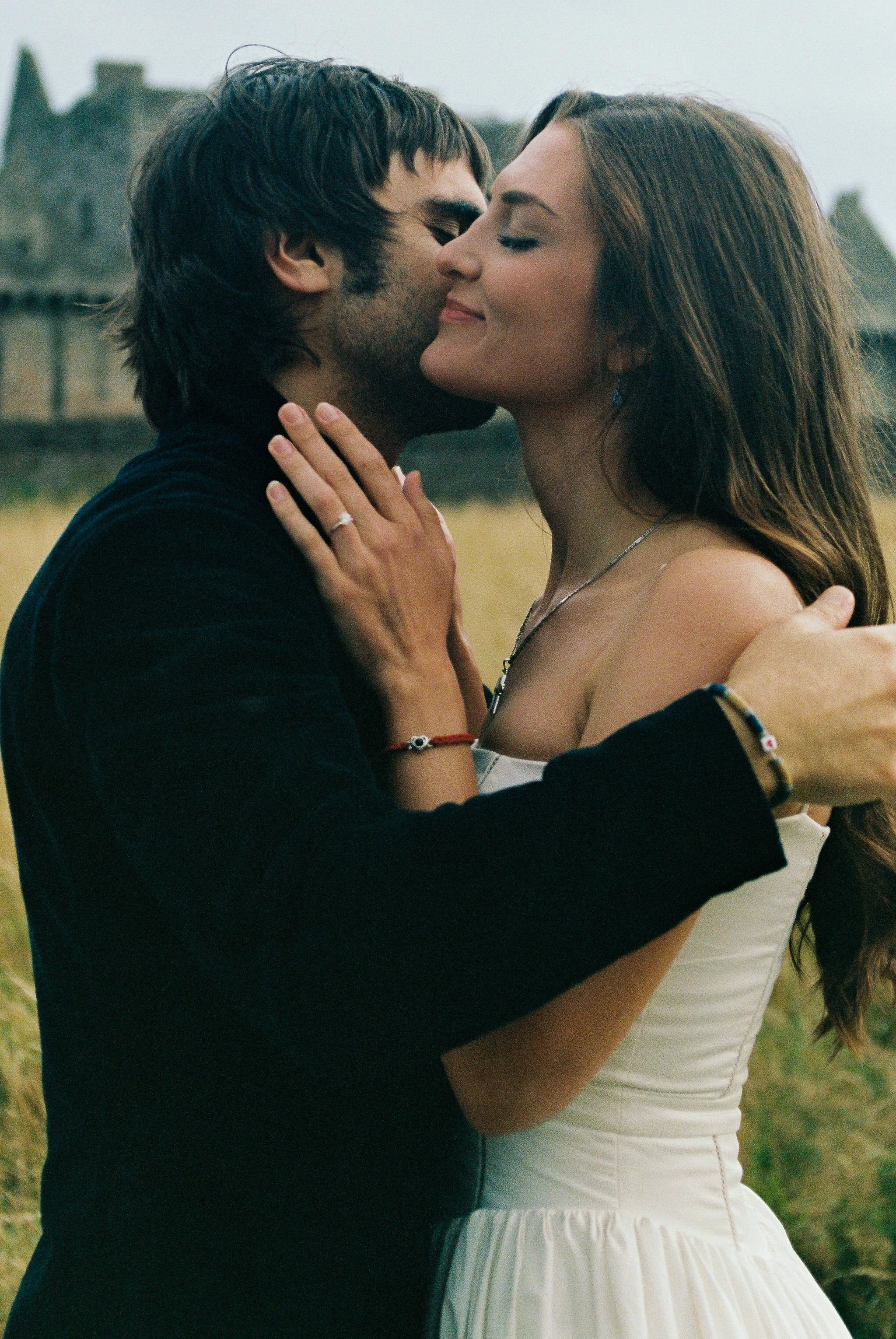 A romantic couple embracing outdoors during daytime, with the man kissing the woman's nose and she smiling with eyes closed, both appearing happy.