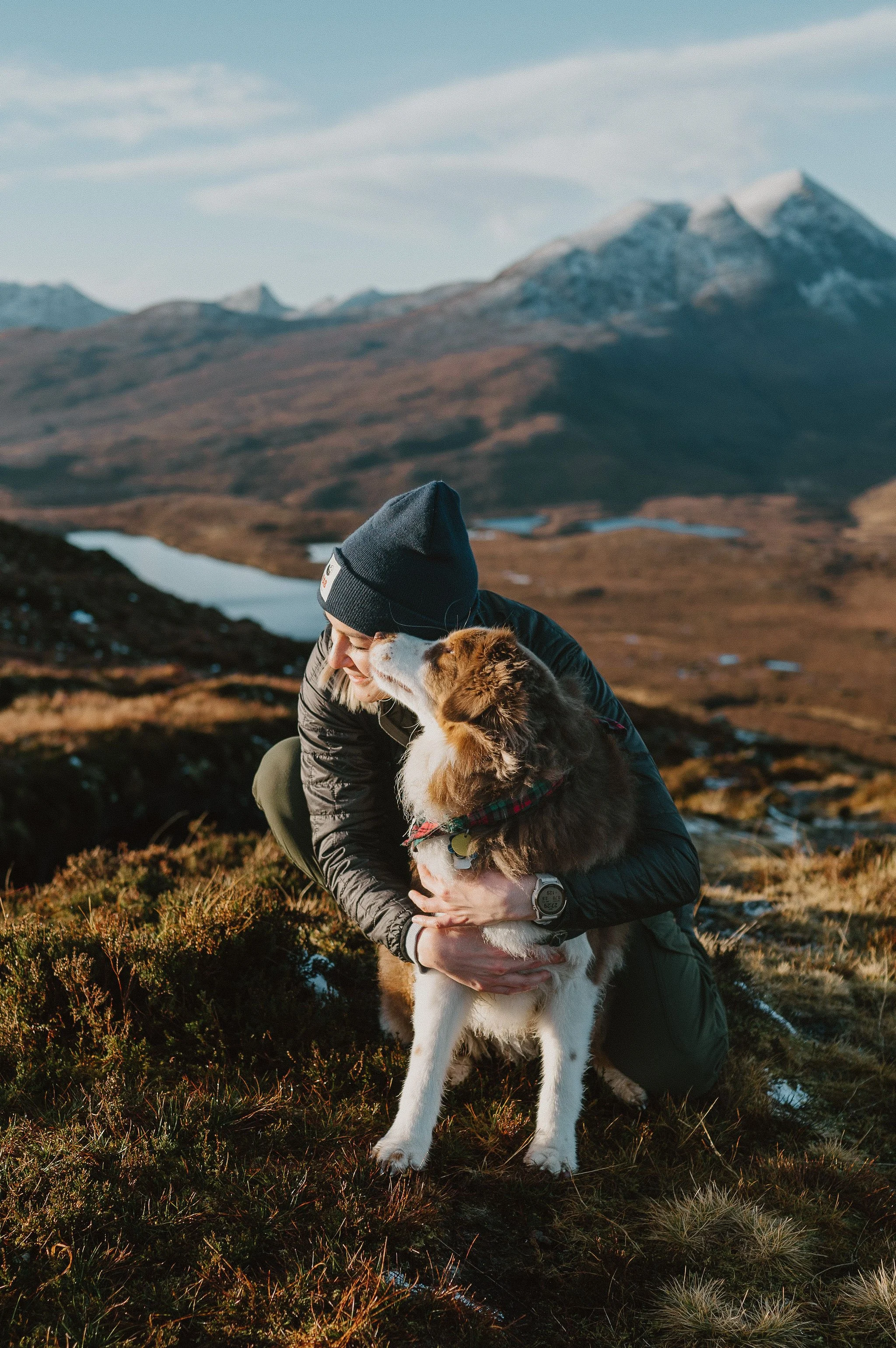 A person wearing a black jacket and gray beanie crouches on a grassy hillside holding a brown and white Australian Shepherd dog. They are outdoors in a mountainous landscape with snow-capped peaks and a lake in the background, during what appears to be late afternoon or early evening.