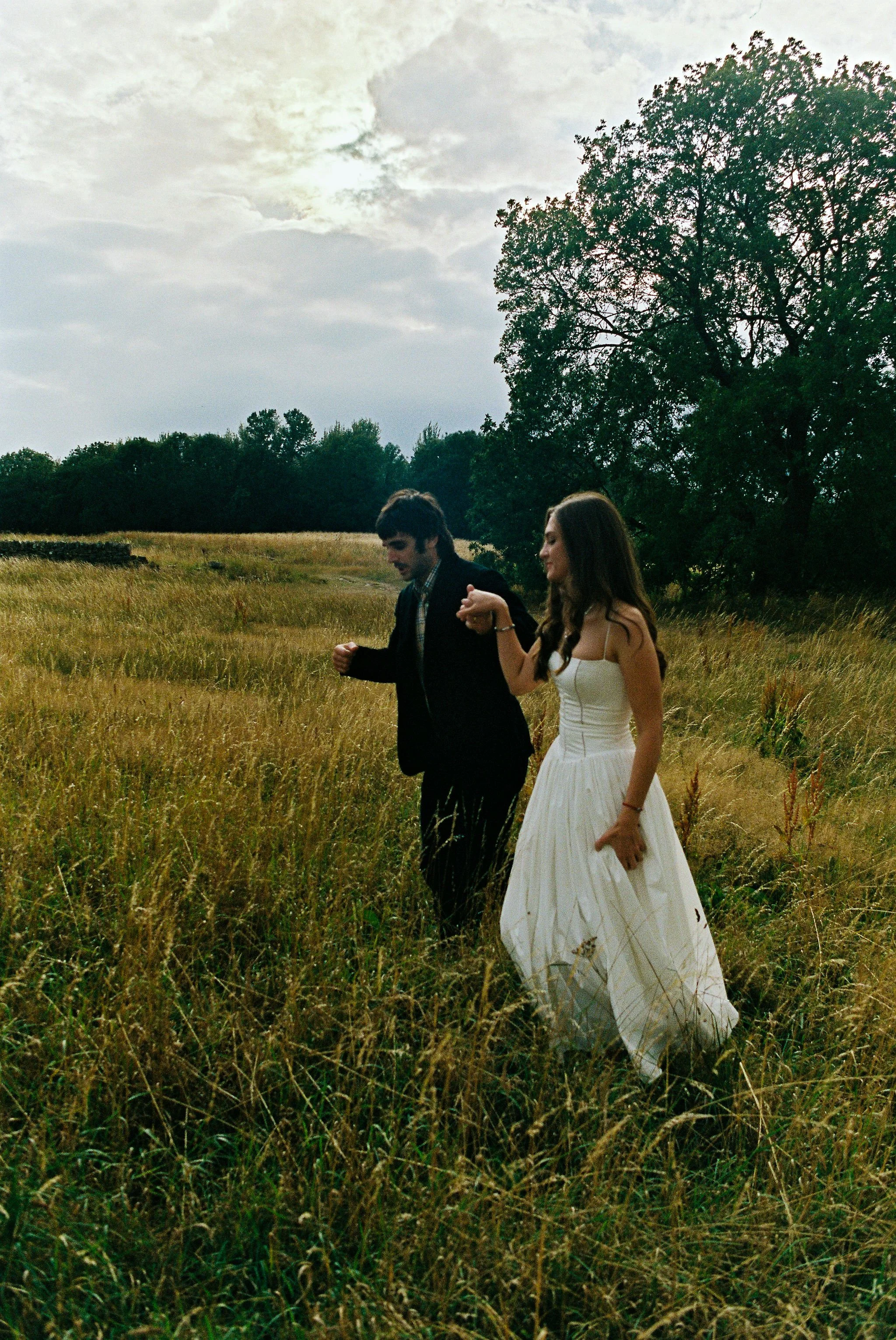 A couple, a man in a dark suit and a woman in a white dress, walking hand-in-hand through a grassy field at sunset.