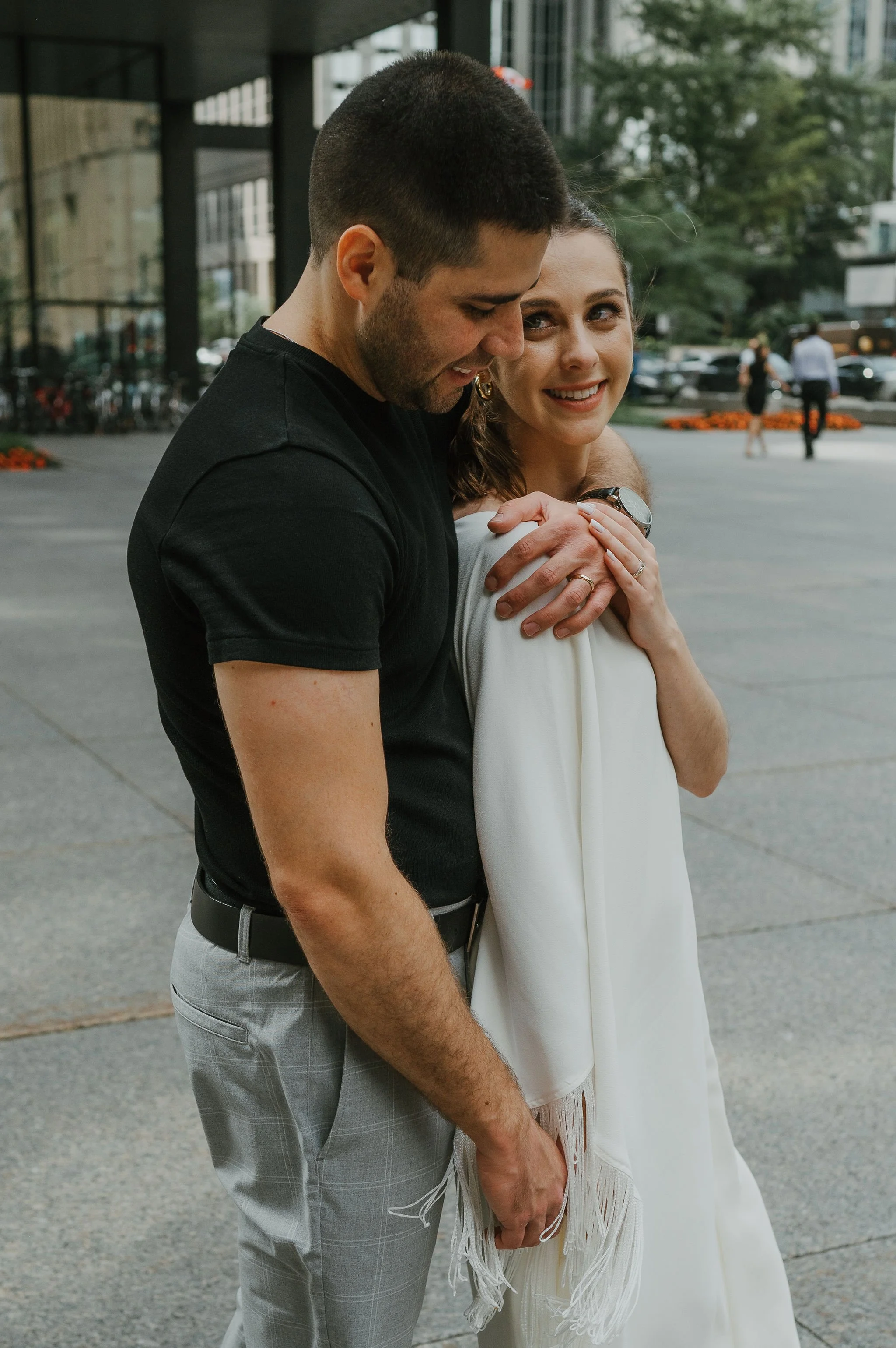 A young couple standing close together outside in a city environment, smiling, with the man embracing the woman from behind.