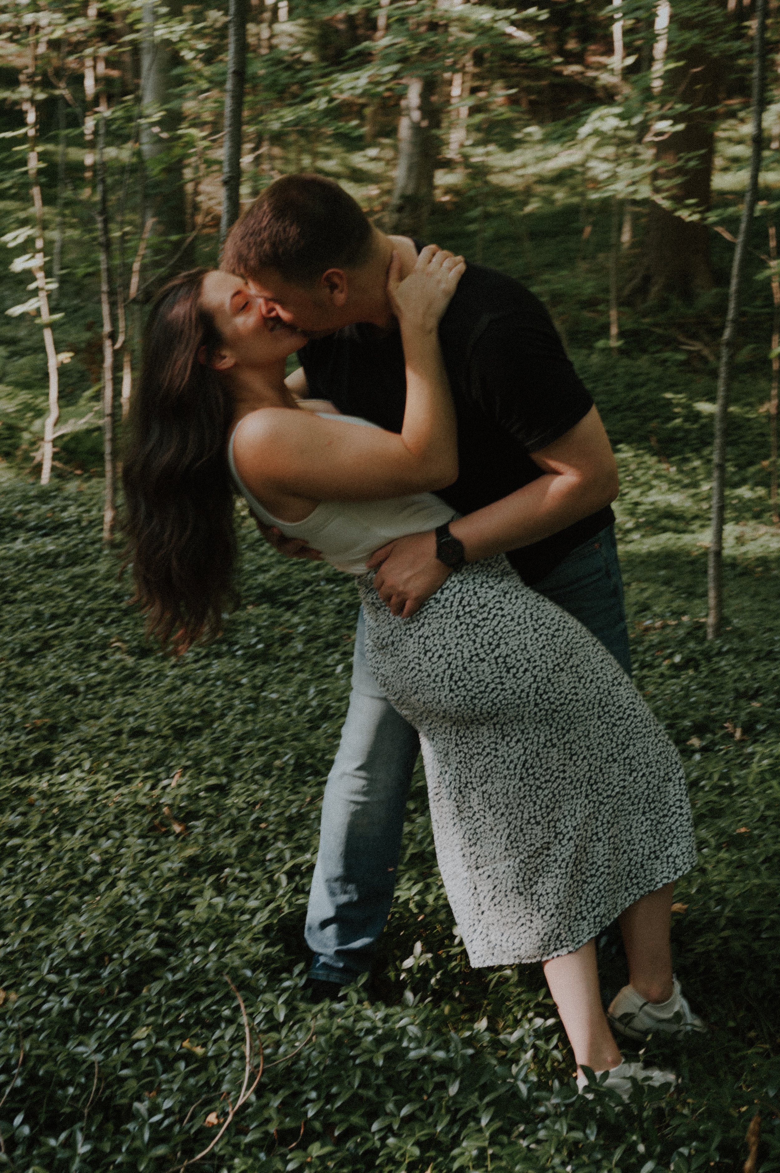 A couple kissing passionately in a forest, with the woman leaning back and the man holding her around the waist.