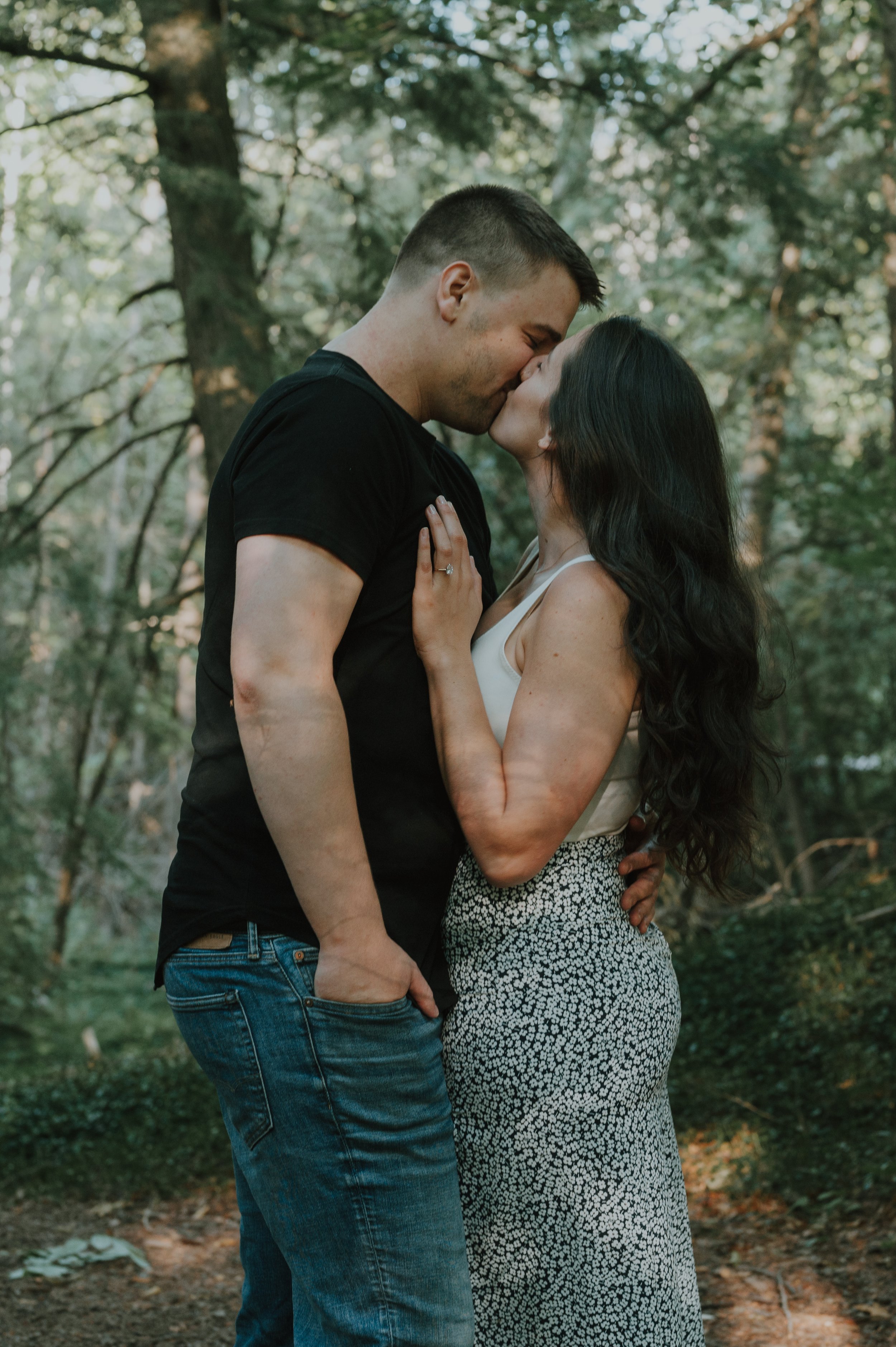 A man and woman kissing outdoors in a wooded area, with the woman holding onto the man's chest and the man with his hand in his pocket.
