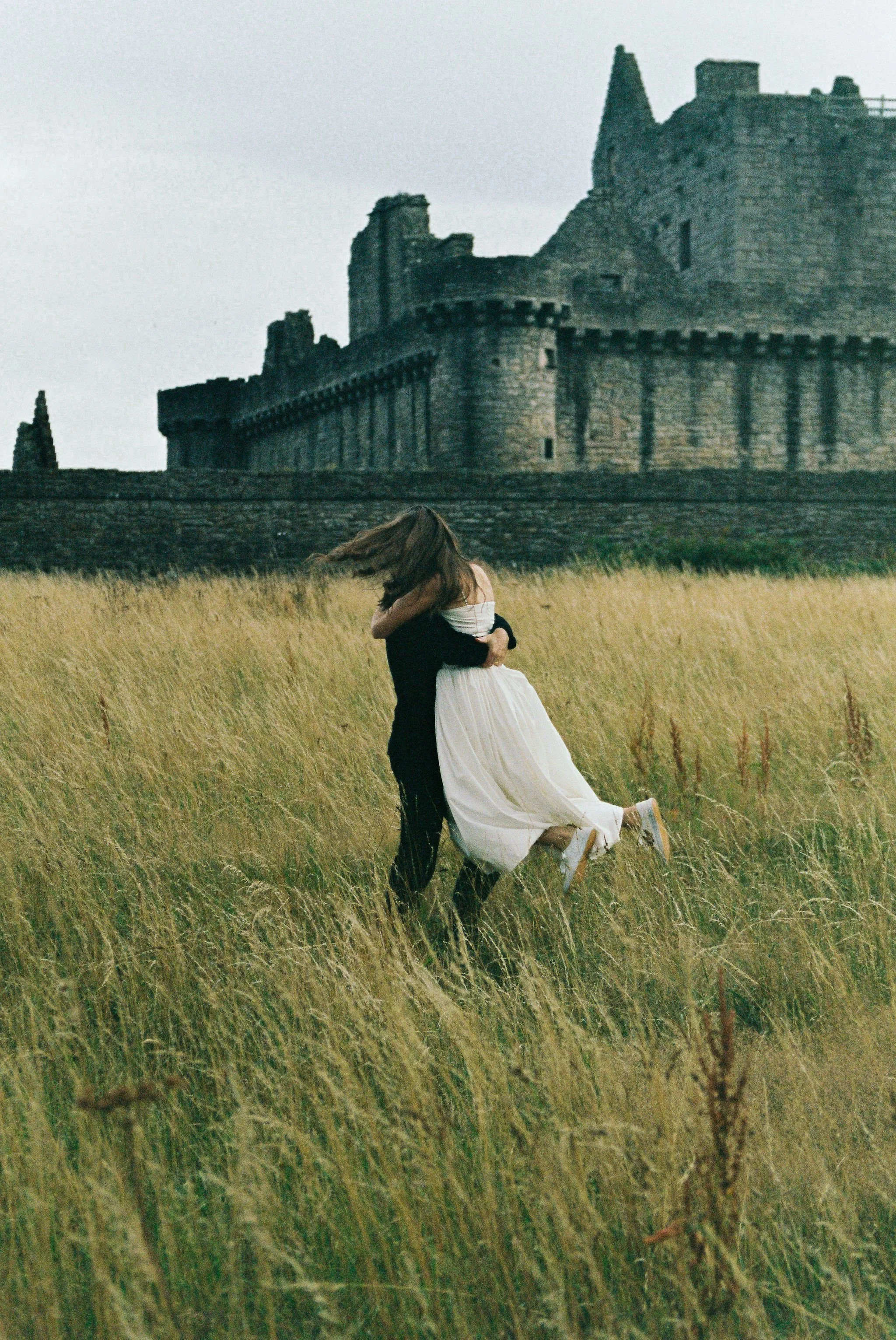 A couple embracing in a field with a castle in the background, the man in black and the woman in a white dress.