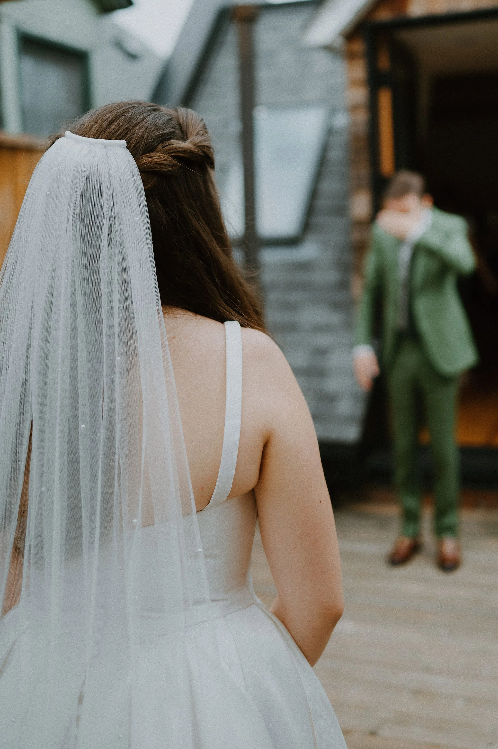 A bride standing outdoors, wearing a white wedding dress and veil, looking at a man in a green suit in the background.