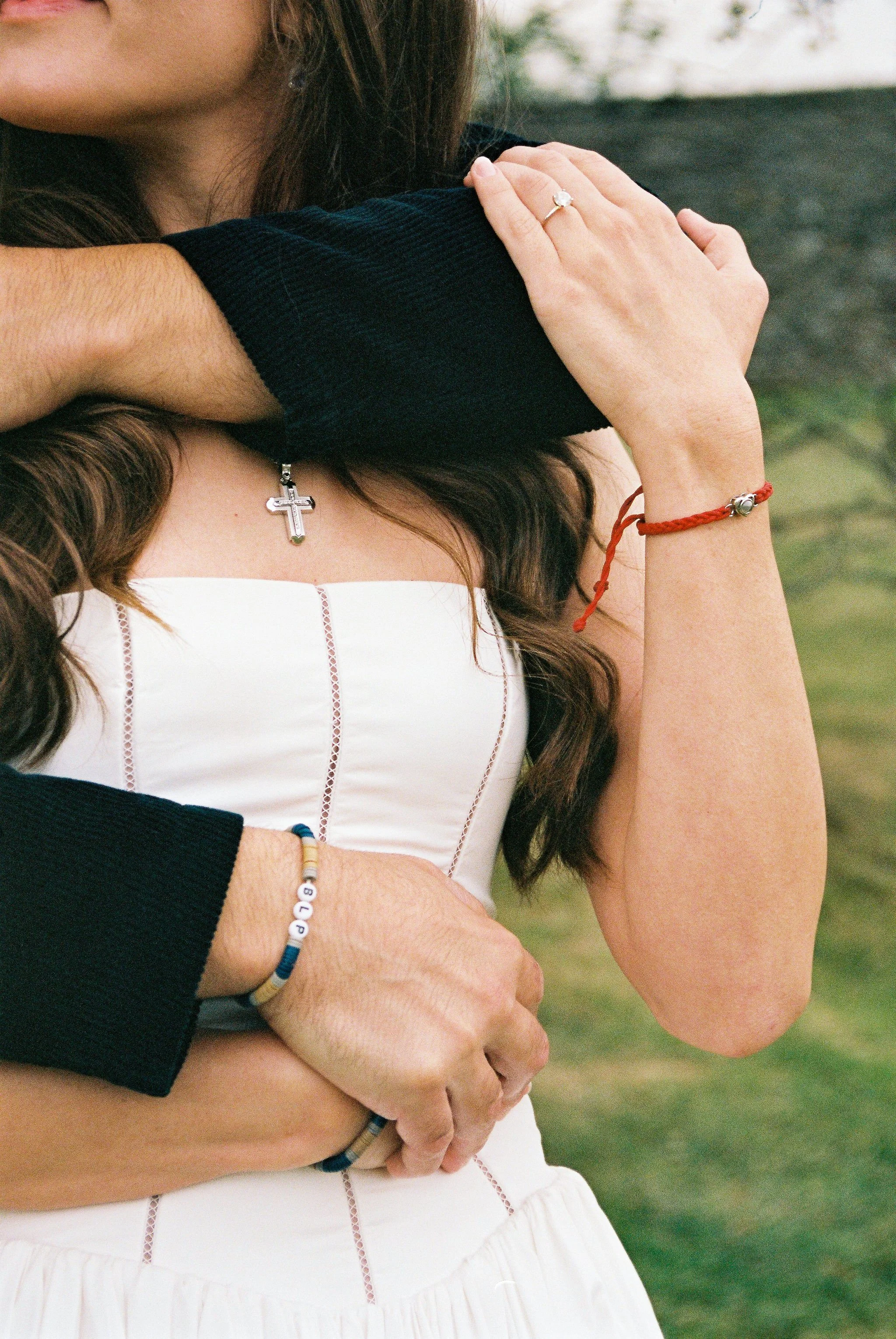 A woman is receiving a hug from another person with an outdoor background. The woman is wearing a white dress with red stitching, a silver cross necklace, and a red bracelet. The other person, dressed in a black top, has their arm around her, showing