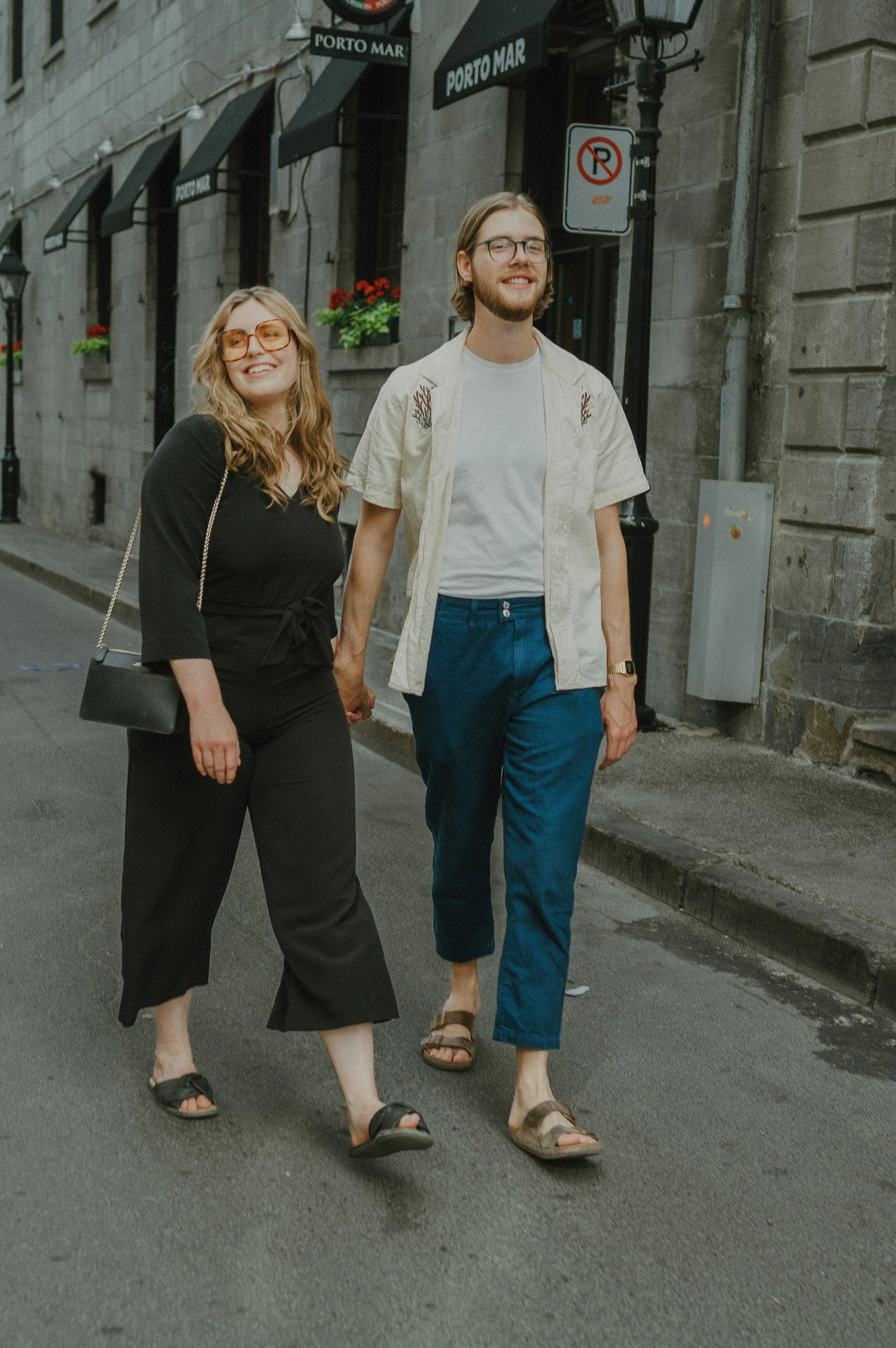A smiling man and woman walking hand in hand on a city street, with storefronts and street signs in the background.