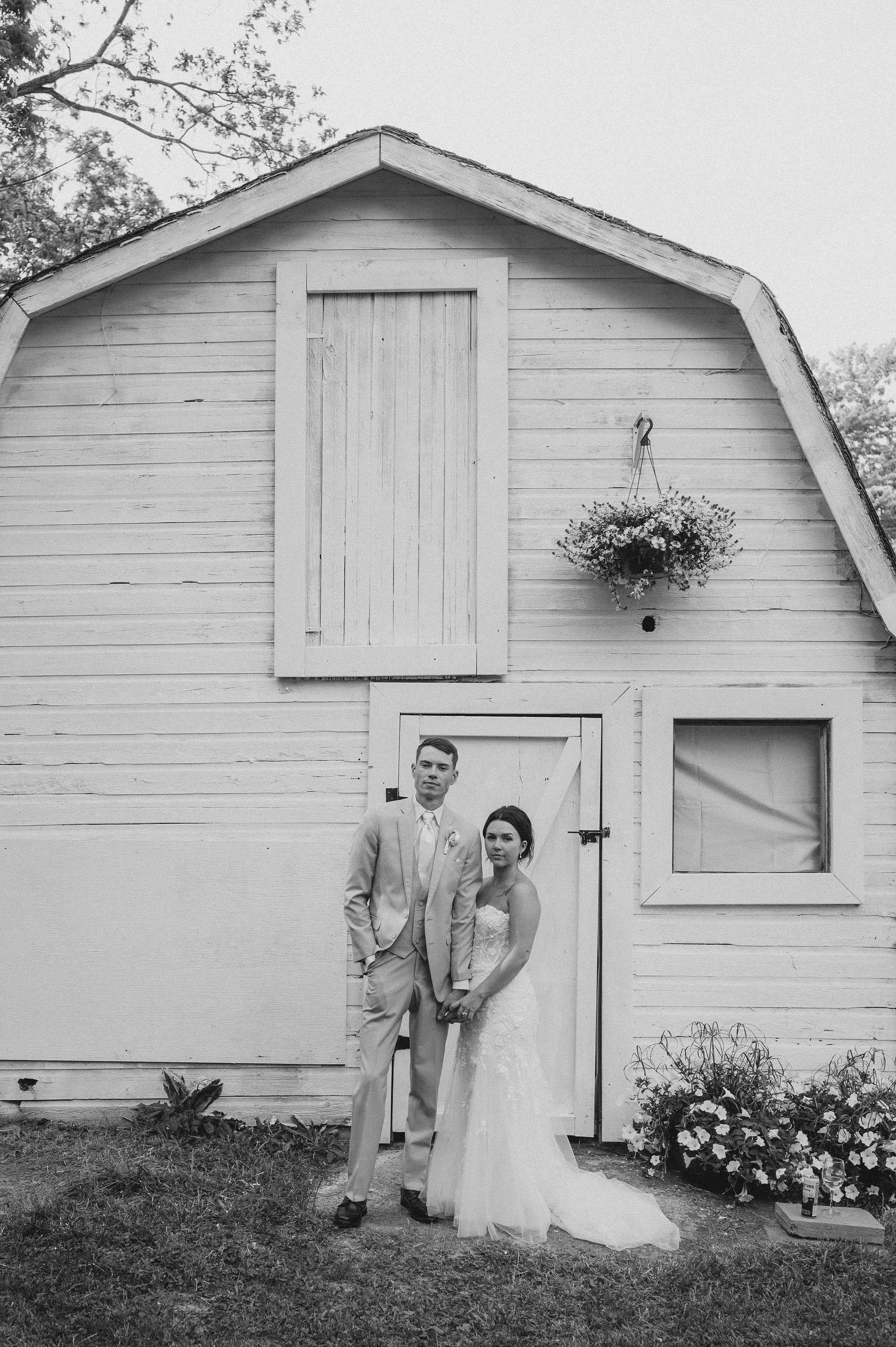 Black and white photo of a bride and groom standing in front of a rustic wooden barn. The groom is wearing a light suit and the bride is in a wedding dress, holding hands. There are potted flowers and plants nearby and a hanging flower basket on the 