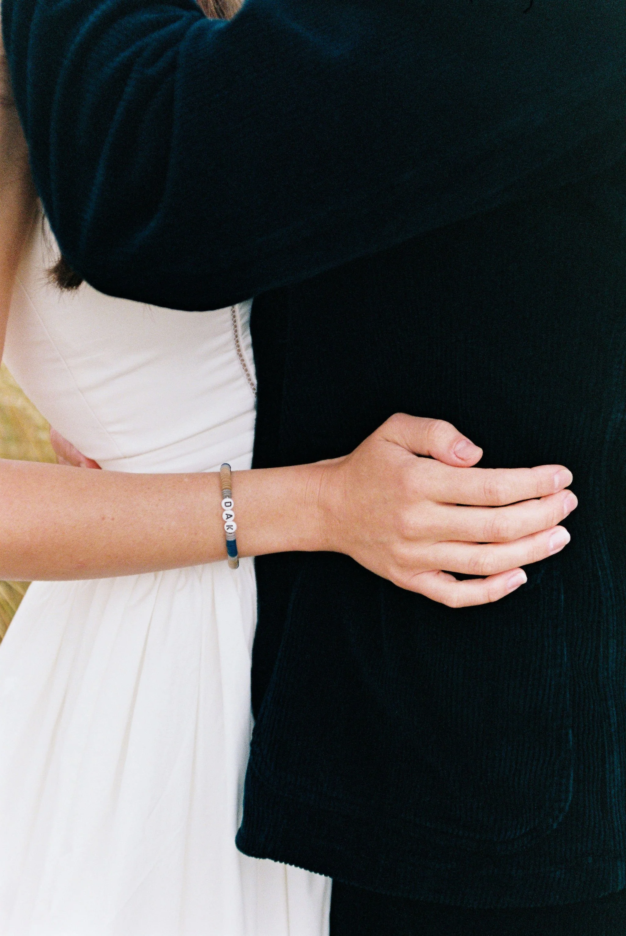 A person in a white dress and a person in black clothing embracing, with the person in white wearing a bracelet that spells 'DAD' on their wrist.