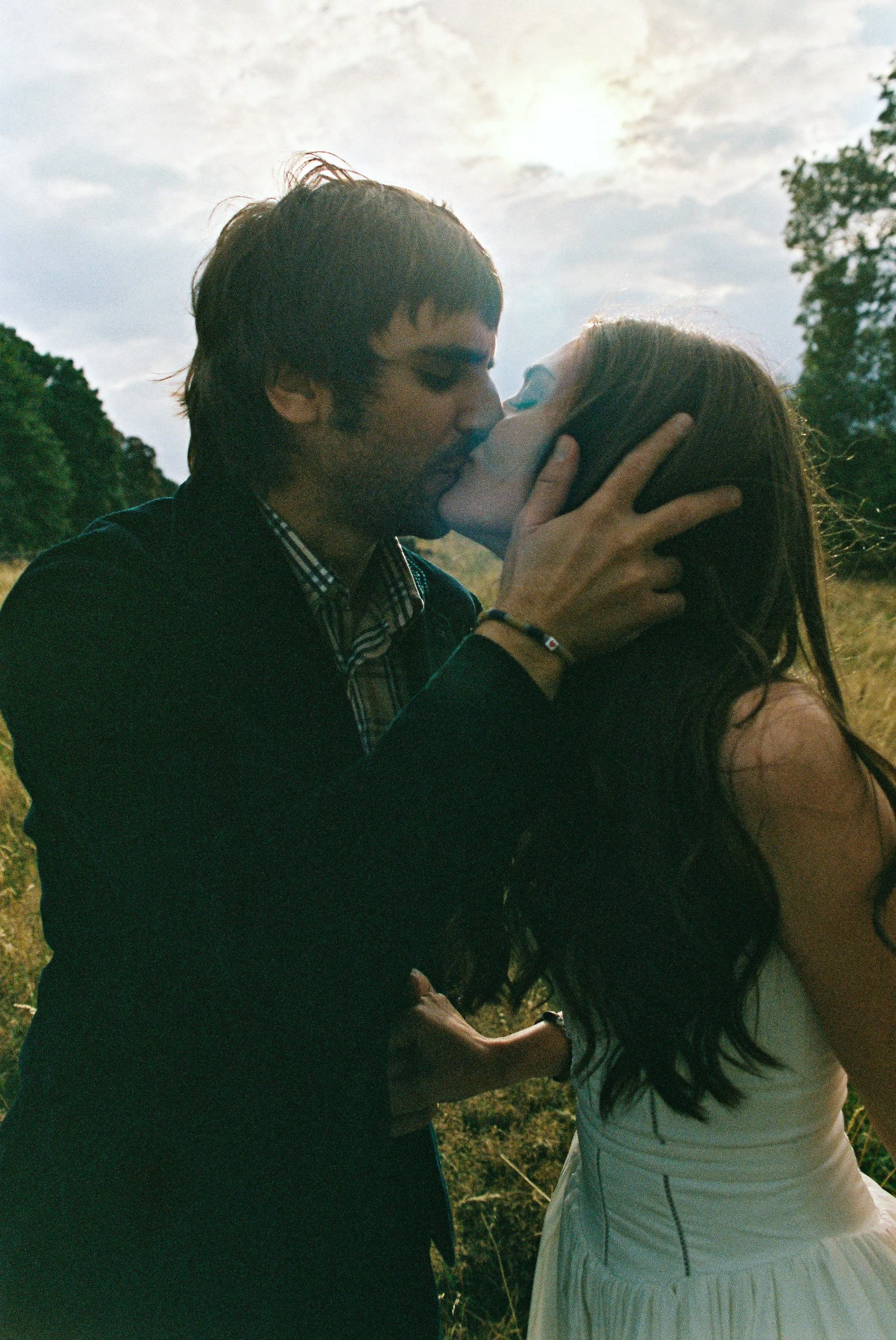 A man and woman kissing outdoors during sunset, with trees and cloudy sky in the background.