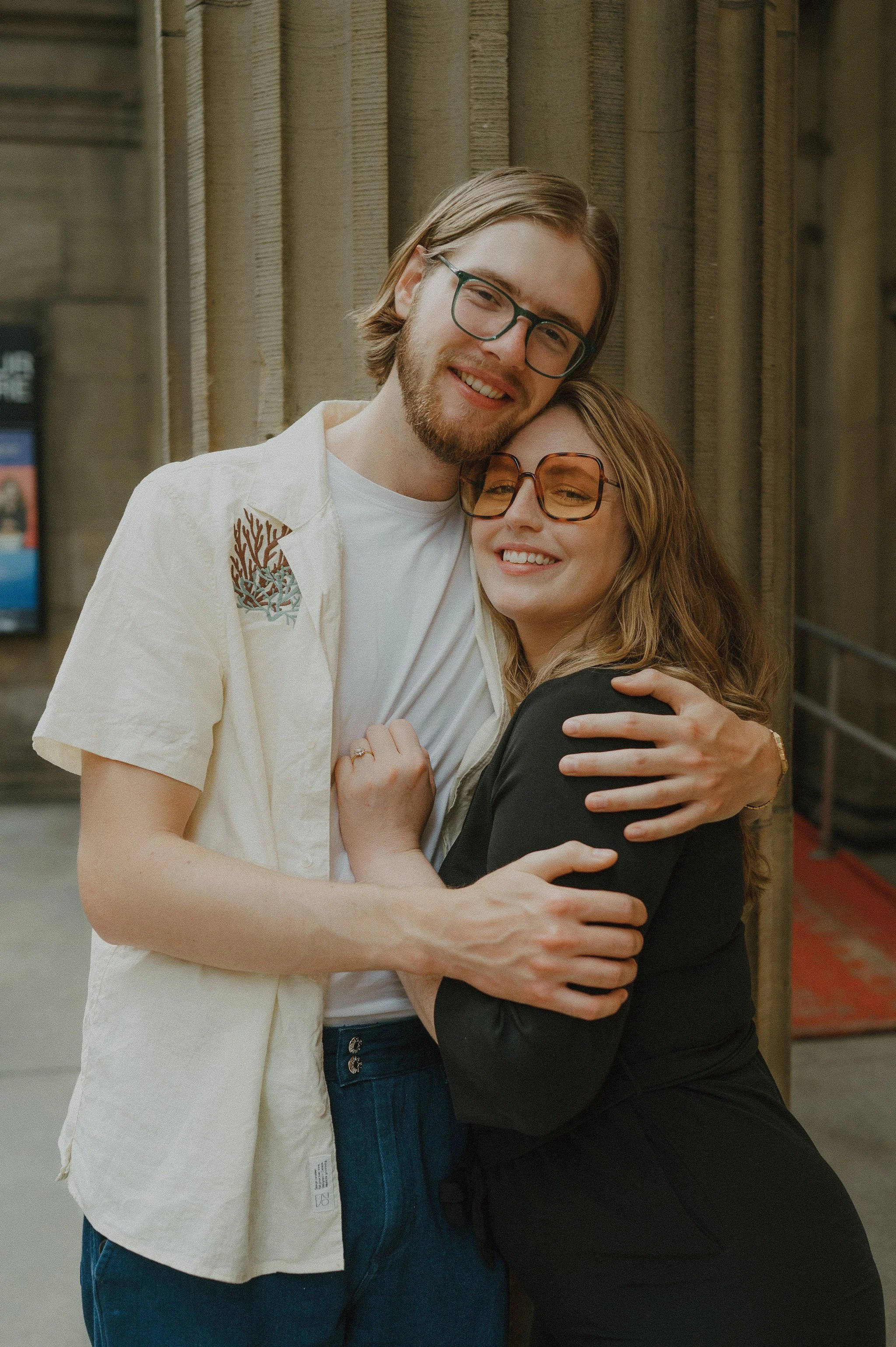 A man and woman are hugging and smiling in front of a brick wall, both wearing glasses.