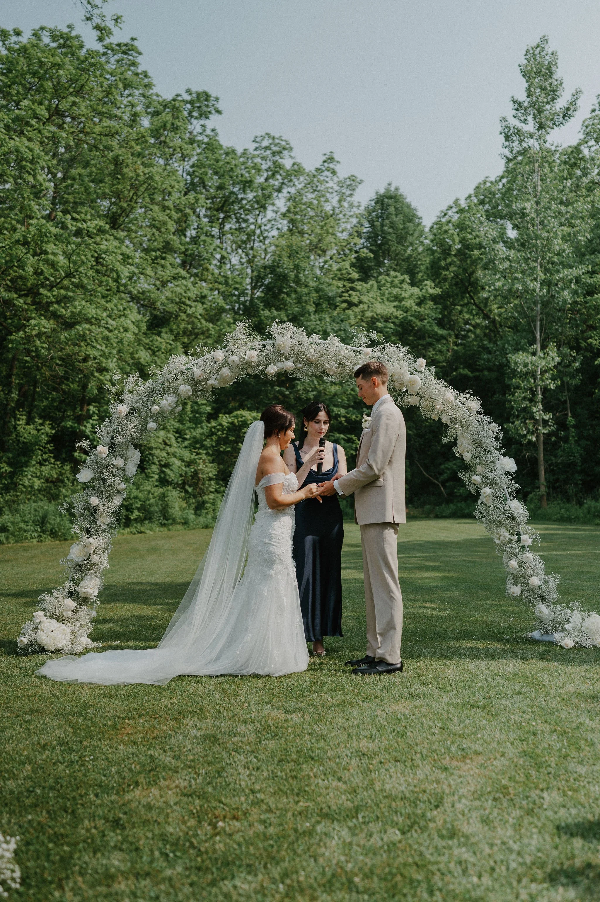 A wedding ceremony outdoors with a bride and groom under a floral arch, officiant, and green trees in the background.