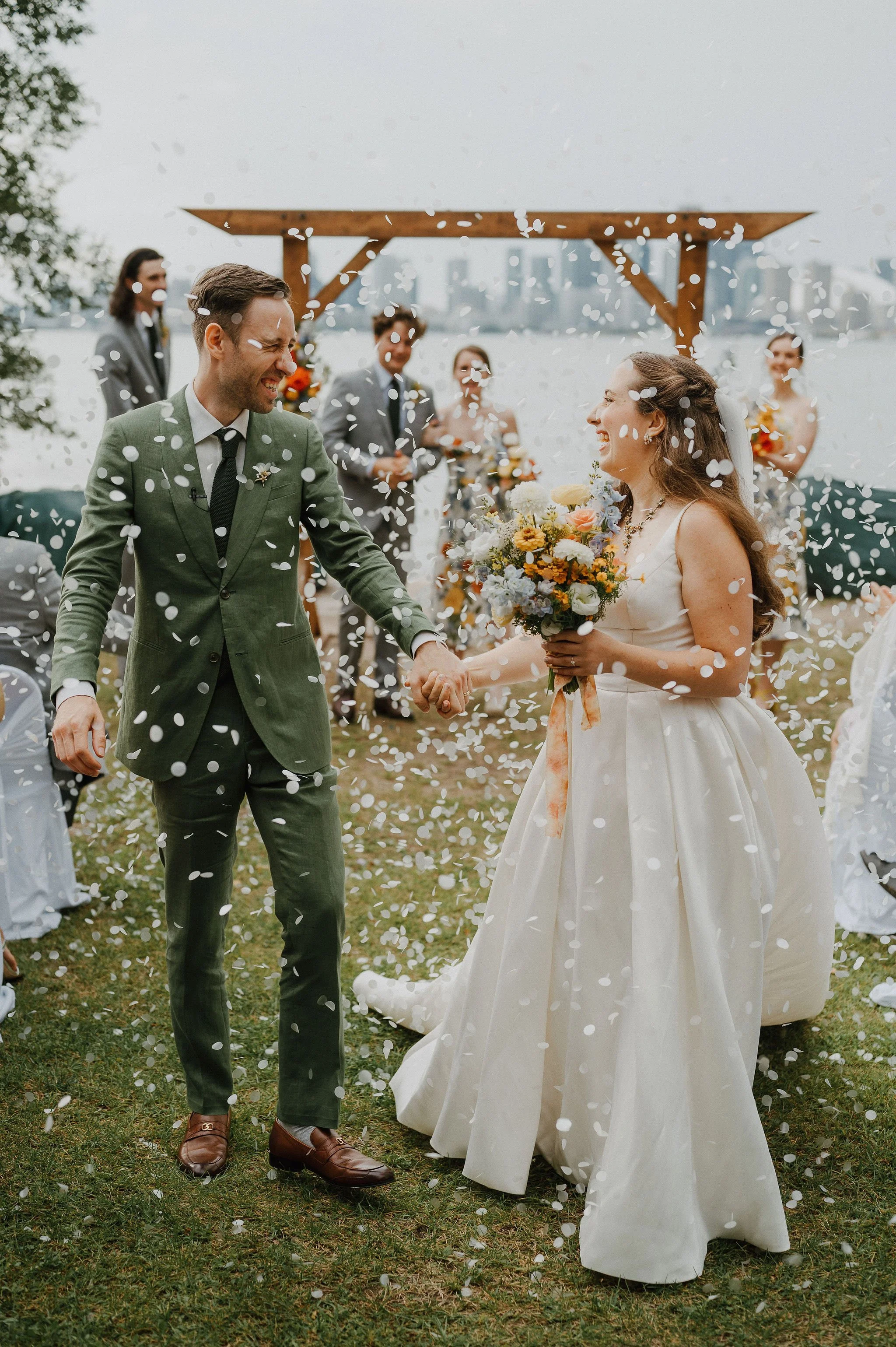 A bride and groom holding hands and smiling at each other during their outdoor wedding ceremony, with guests and a wooden arch in the background, while white confetti falls around them.