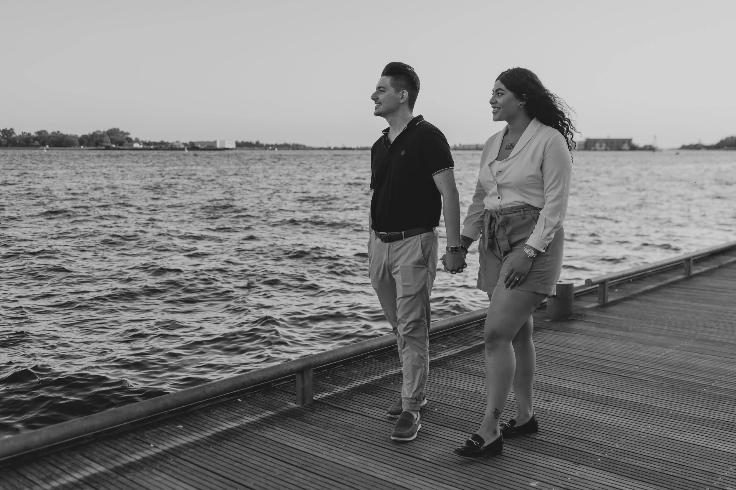 A smiling couple walking hand in hand along a wooden pier near a body of water during sunset, holding hands and enjoying each other's company.