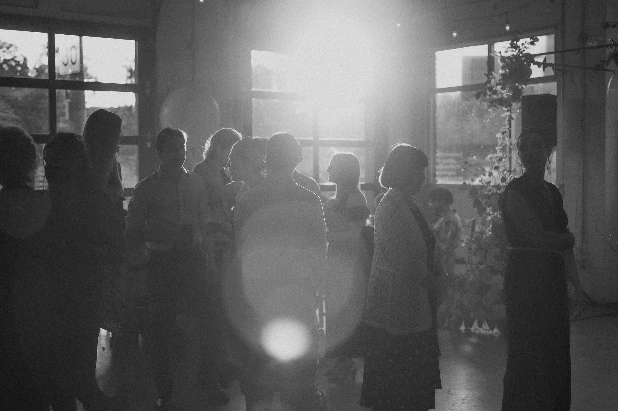 Silhouettes of people socializing inside a room with large windows, sunlight streaming through, and some plants visible.