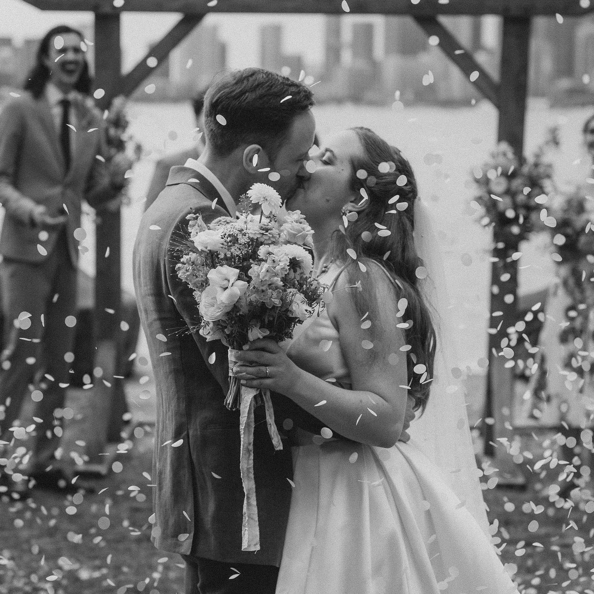Black and white photo of a wedding couple kissing, with the bride holding a bouquet, surrounded by falling confetti, and to the side a person in a suit and glasses smiling.