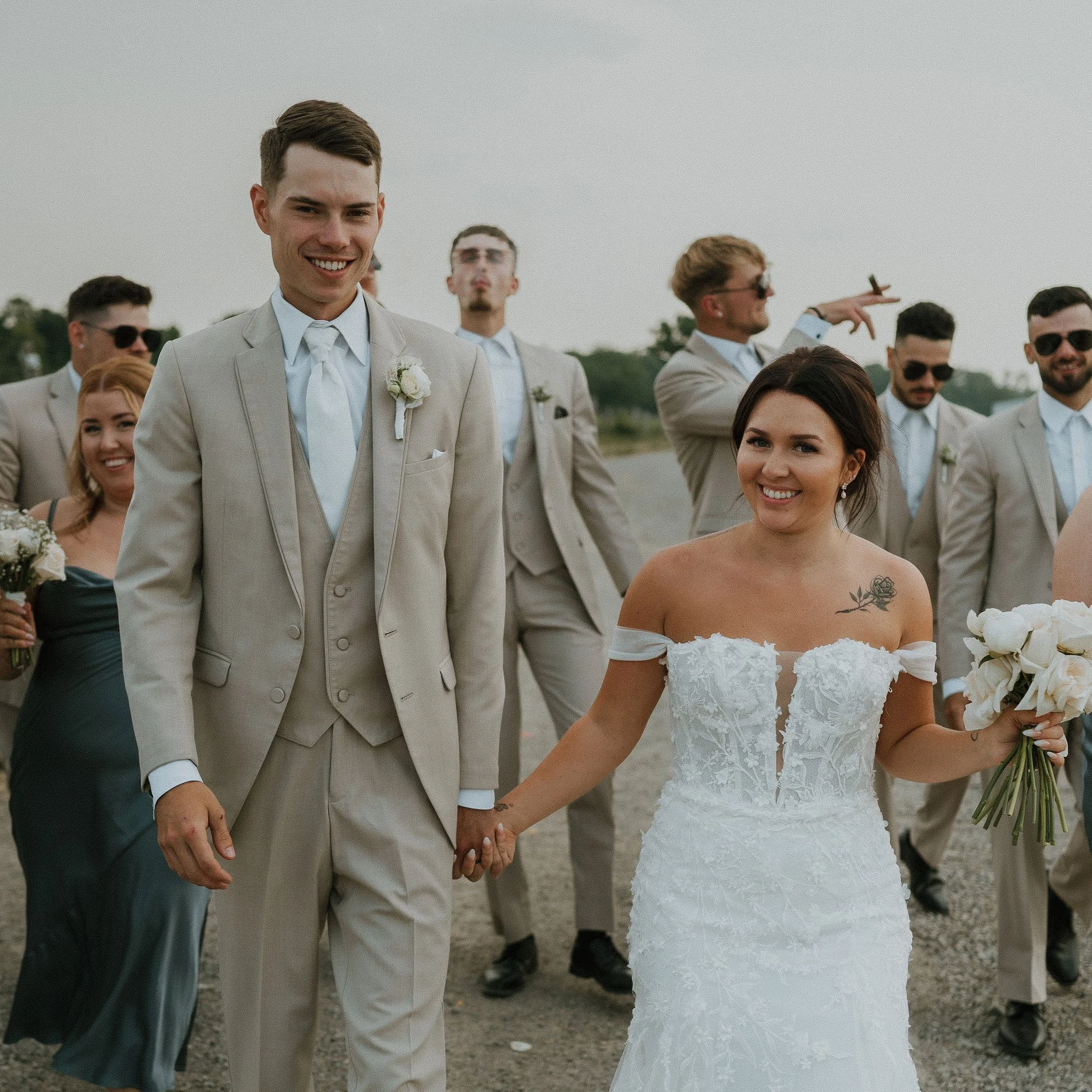 A happy newlywed couple holding hands and walking outdoors with friends, bride in a white lace wedding dress and groom in a beige suit, during a wedding celebration.