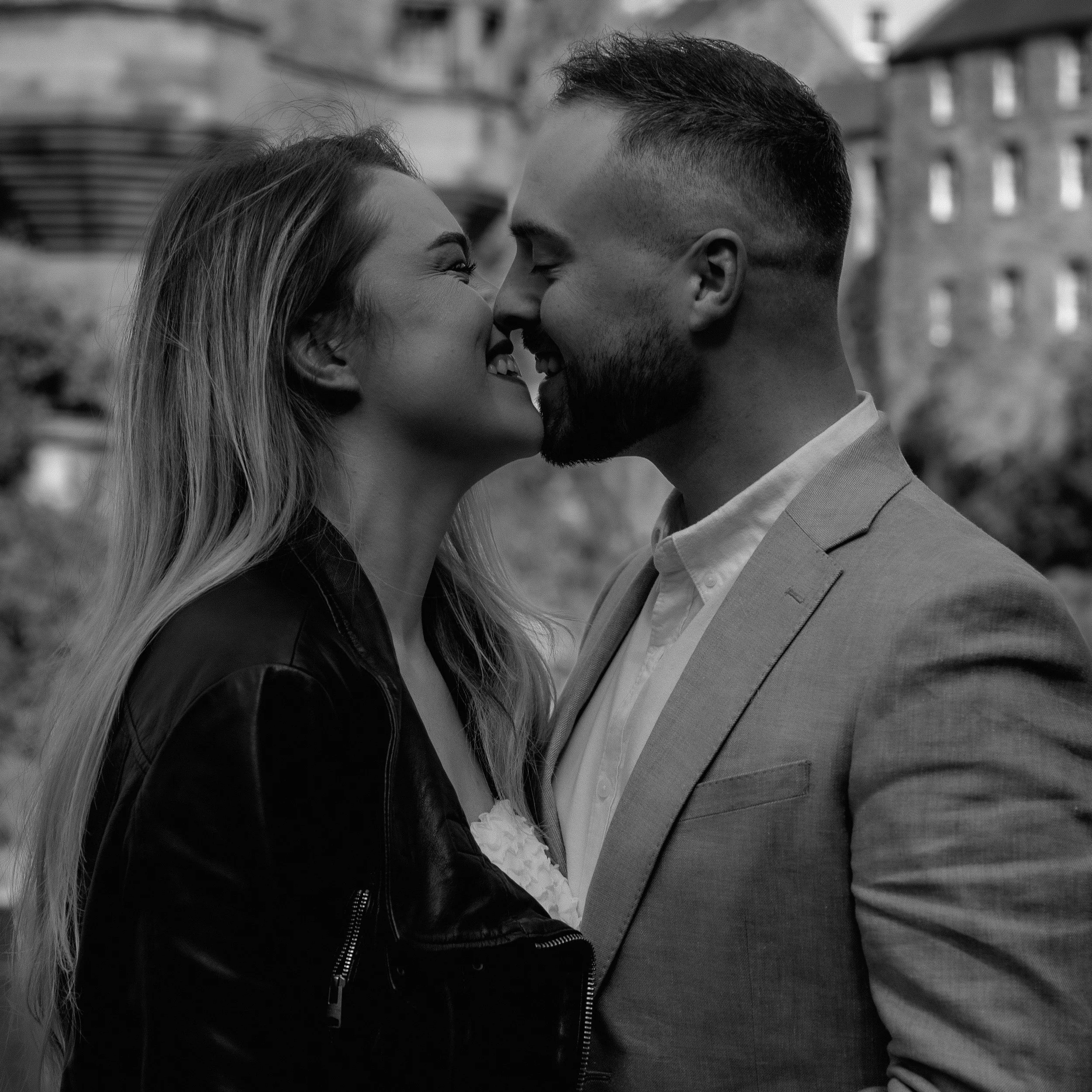 A black and white photo of a couple touching noses and smiling at each other outdoors, with buildings and trees in the background.