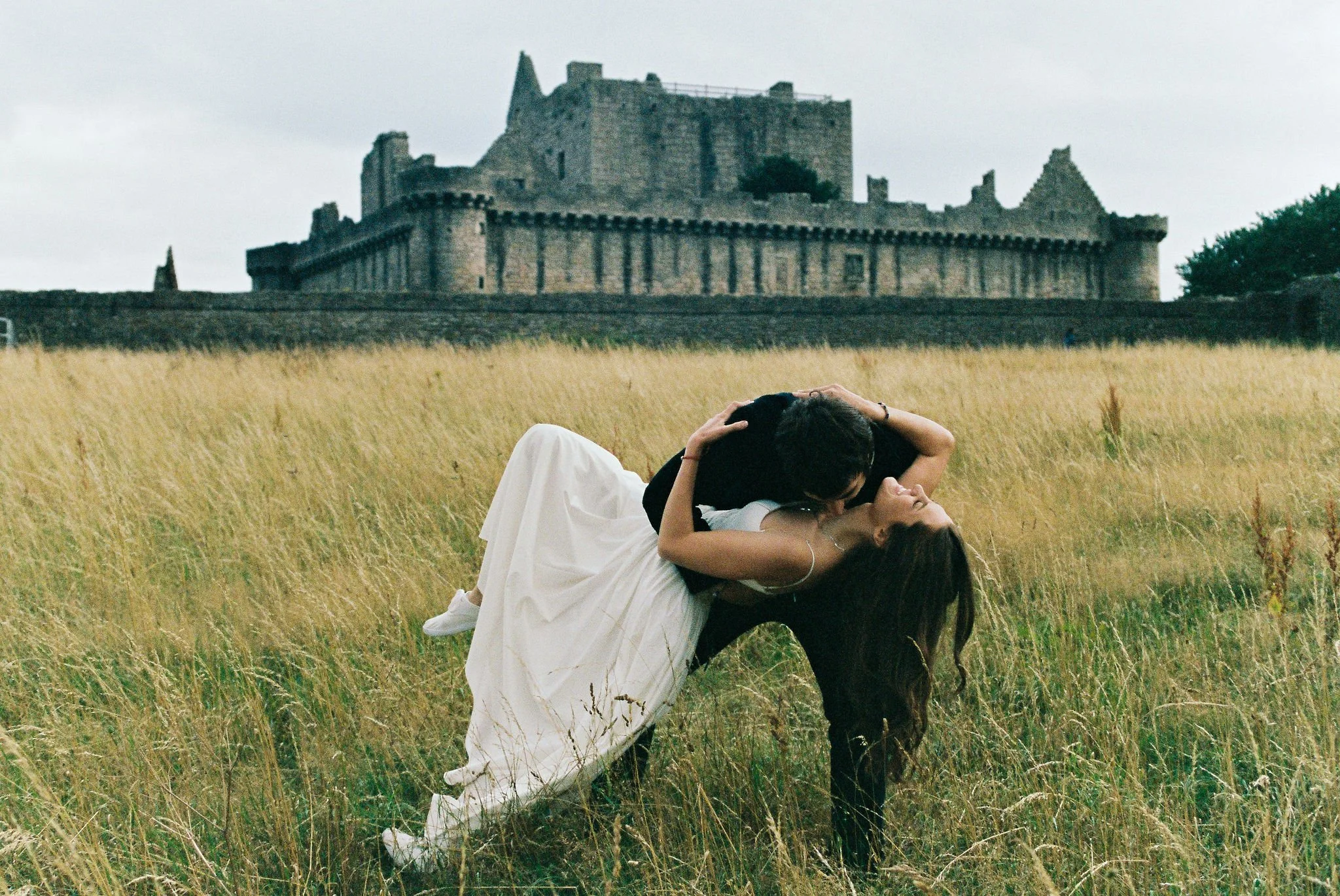 A couple in wedding attire sharing a kiss in a field of tall grass, with a historic stone castle in the background on a cloudy day.