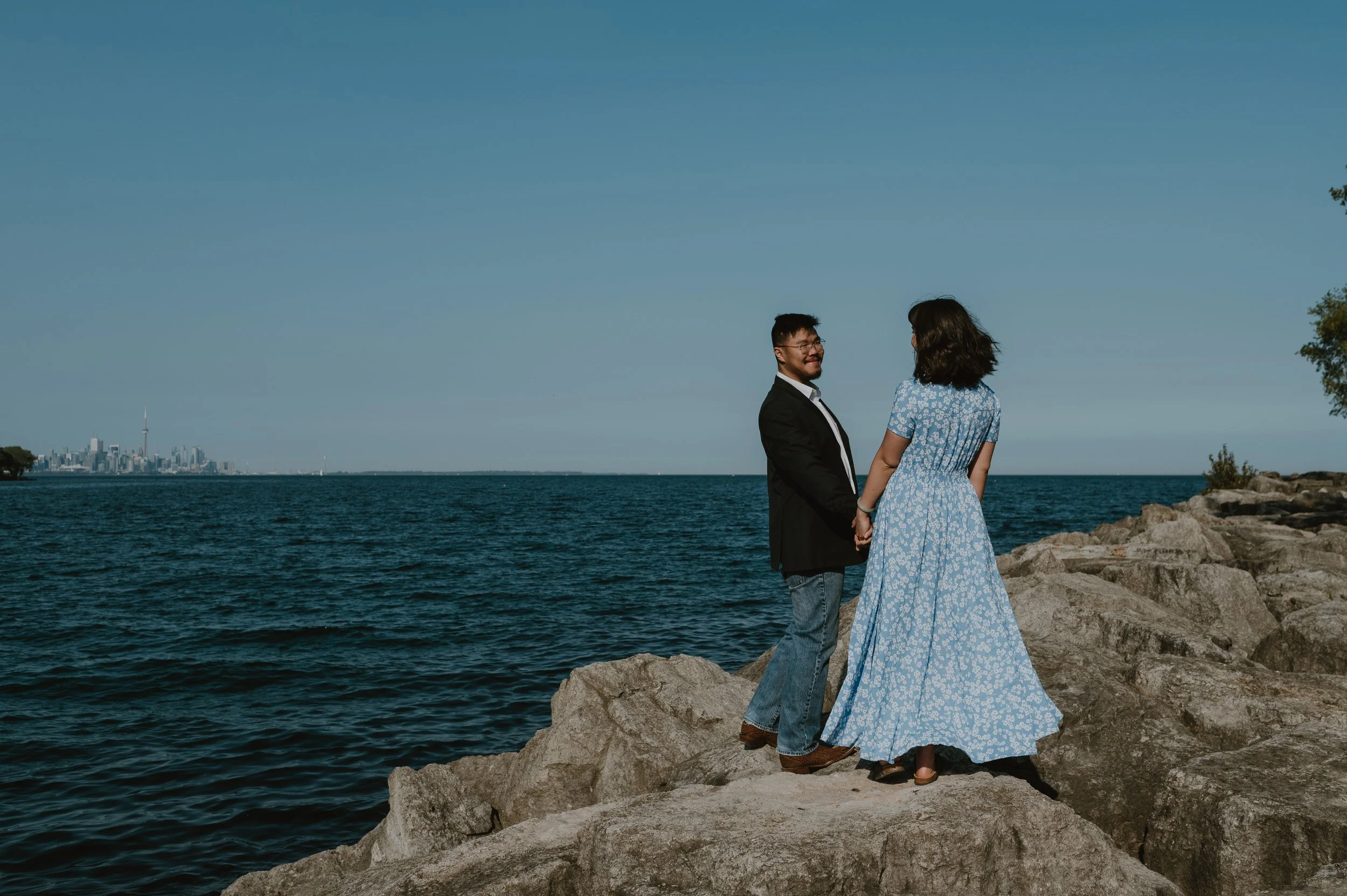 A man and woman standing hand in hand on rocks by a large body of water, with a city skyline in the distance under a clear blue sky.