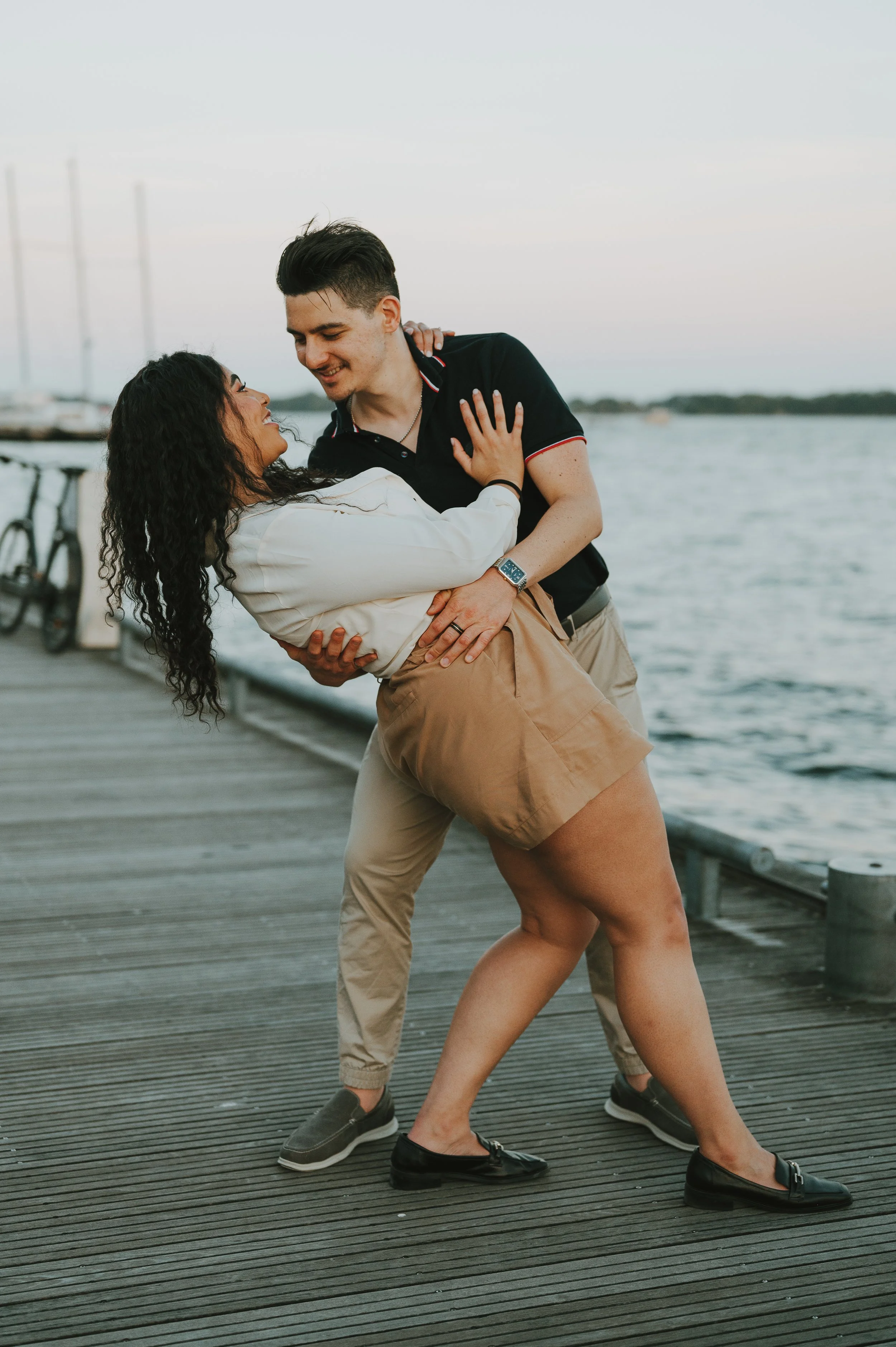 A man and woman dancing on a wooden dock by the water during sunset, smiling and looking at each other.