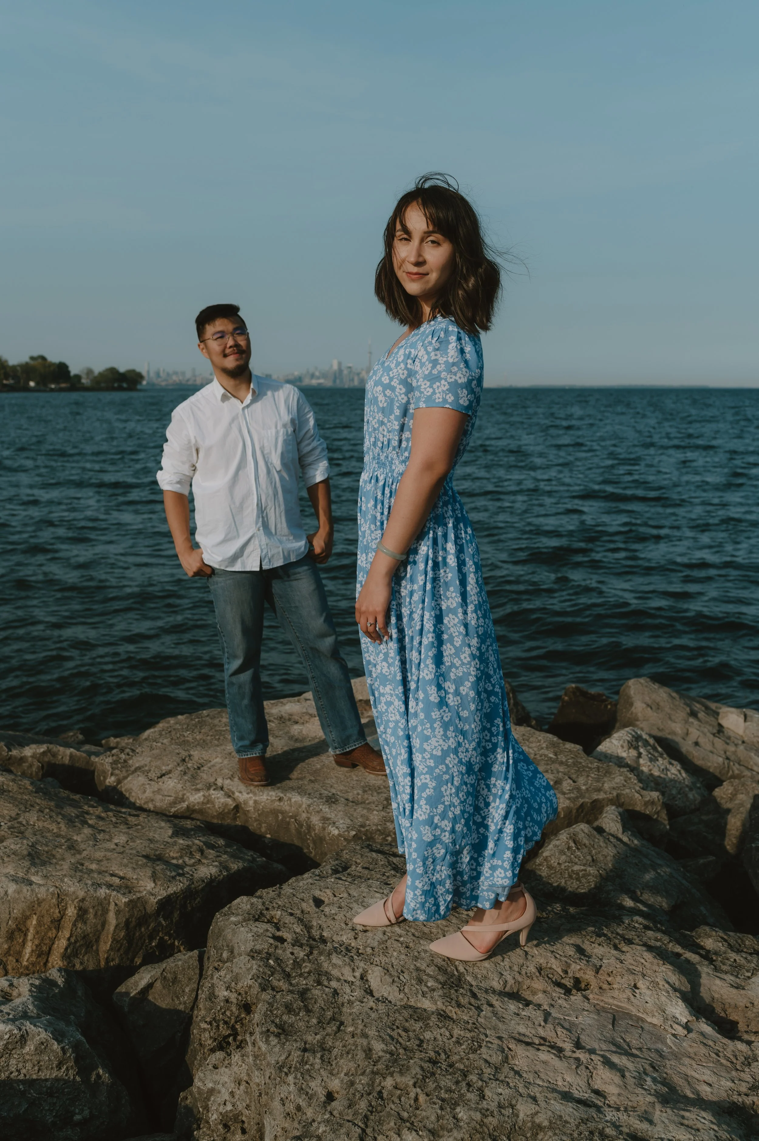 A woman in a blue floral dress and heels standing on rocks by the water, with a man in a white shirt and jeans behind her, at the shoreline with a city skyline in the background.