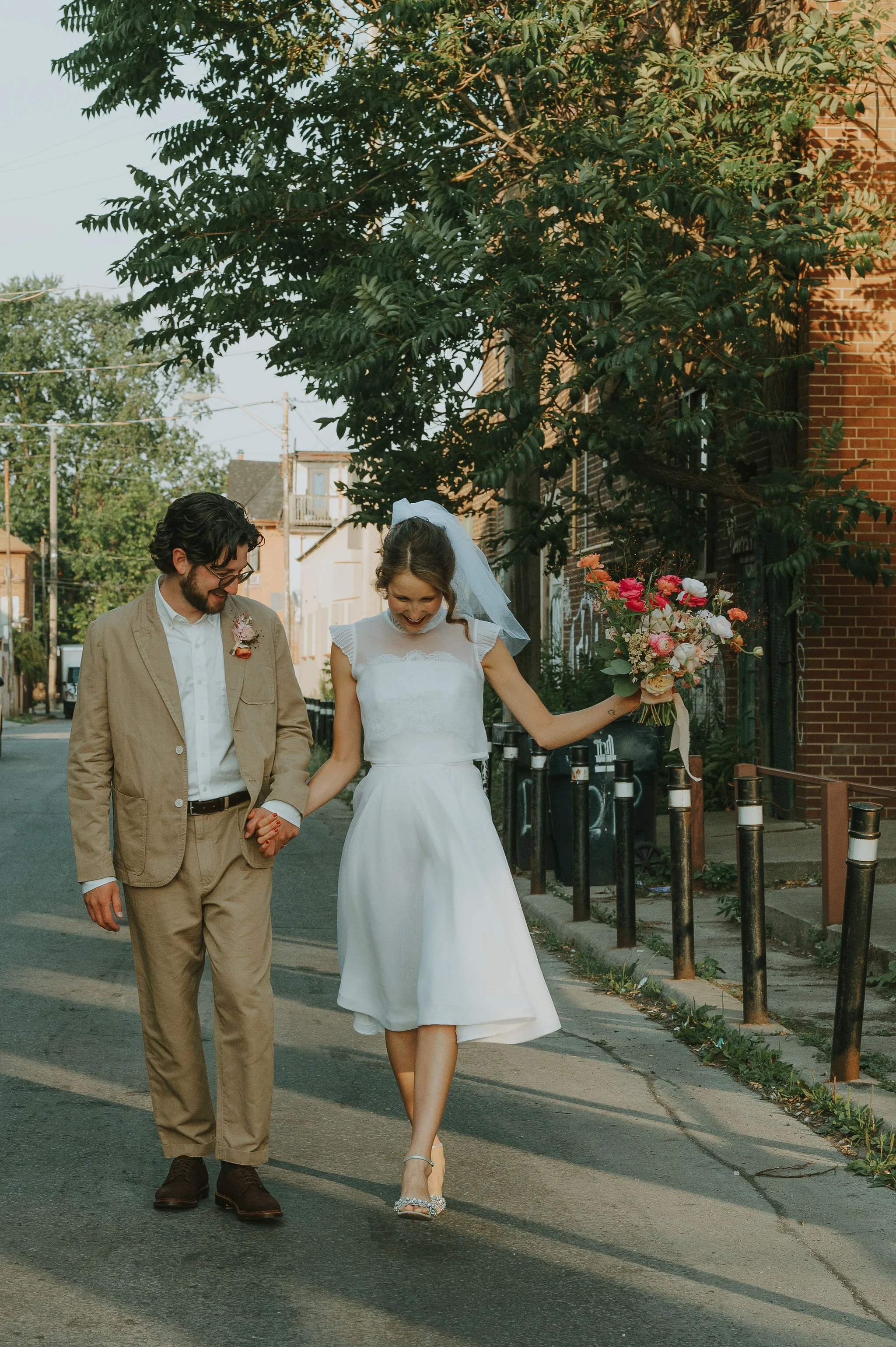 A couple walking hand-in-hand down a street. The woman is wearing a white dress and holding a bouquet of flowers, while the man is dressed in a beige suit. They are smiling and looking at each other.