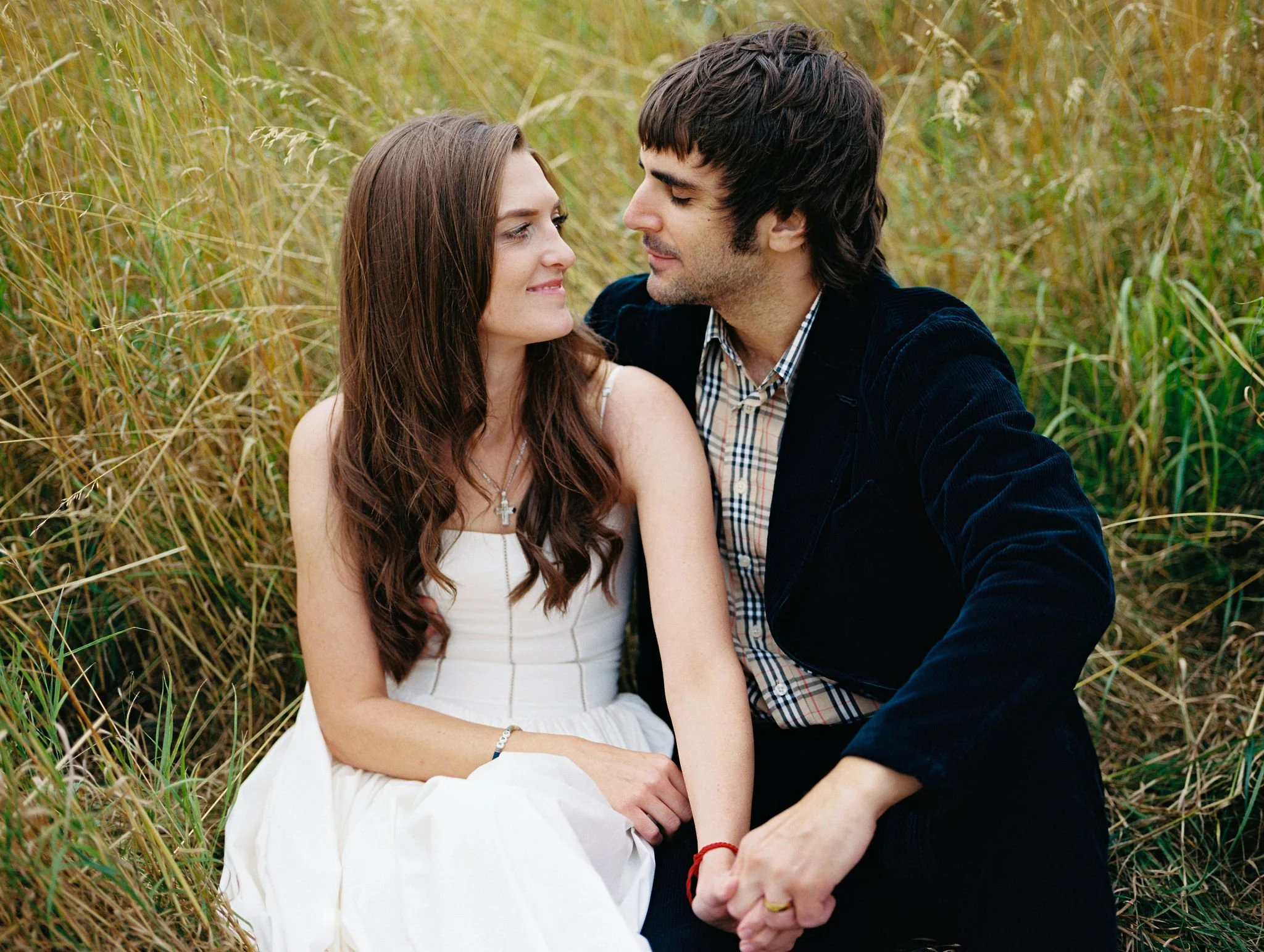 A young couple sitting in a a field of tall grass, looking into each other's eyes, smiling.
