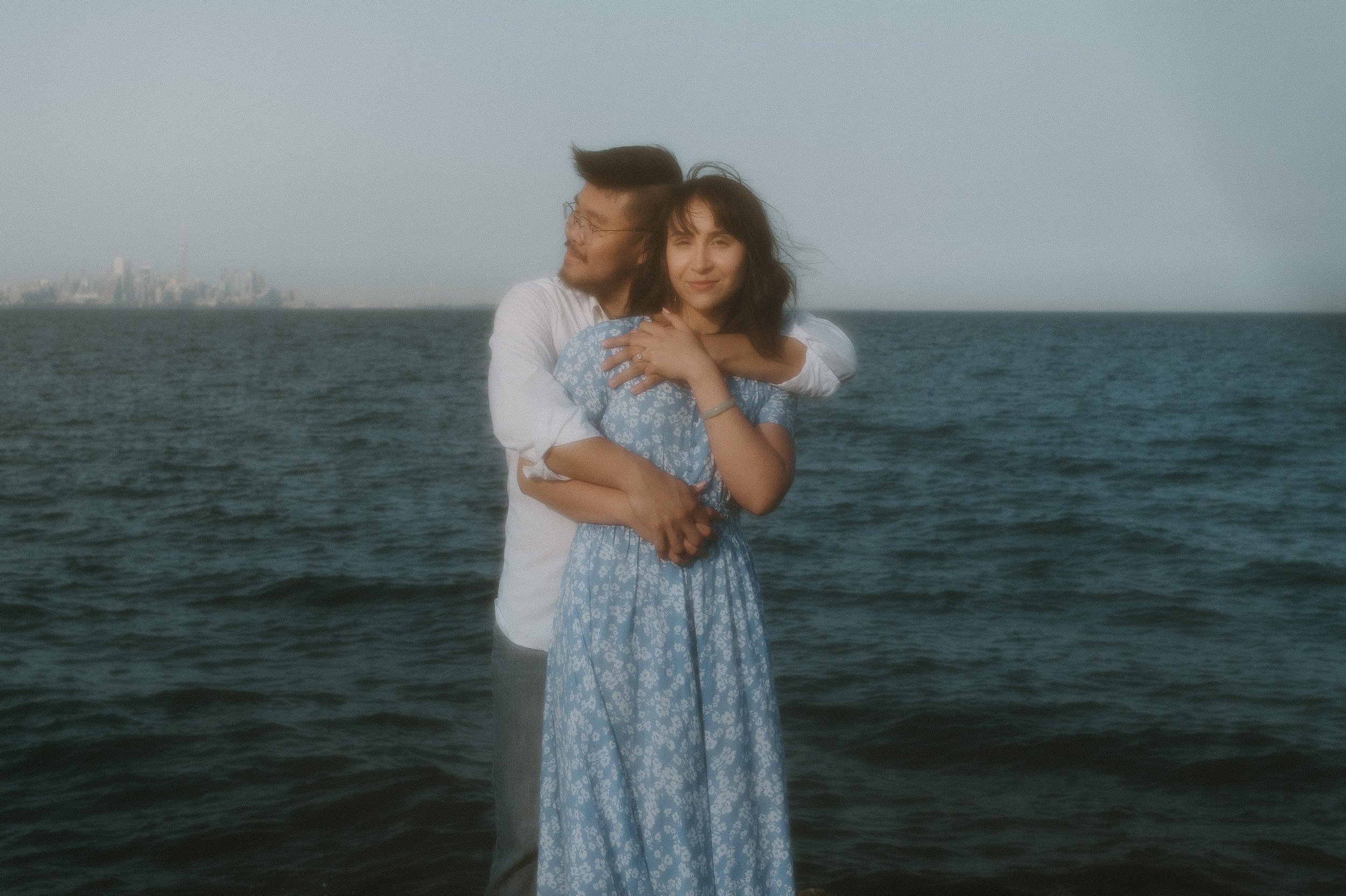 A couple hugging near the ocean with a city skyline in the distance, one woman and one man, both dressed casually.