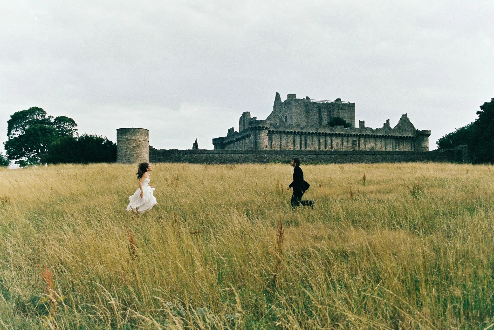 A woman in a white dress running across a grassy field towards a man in black clothing, with a castle in the background.
