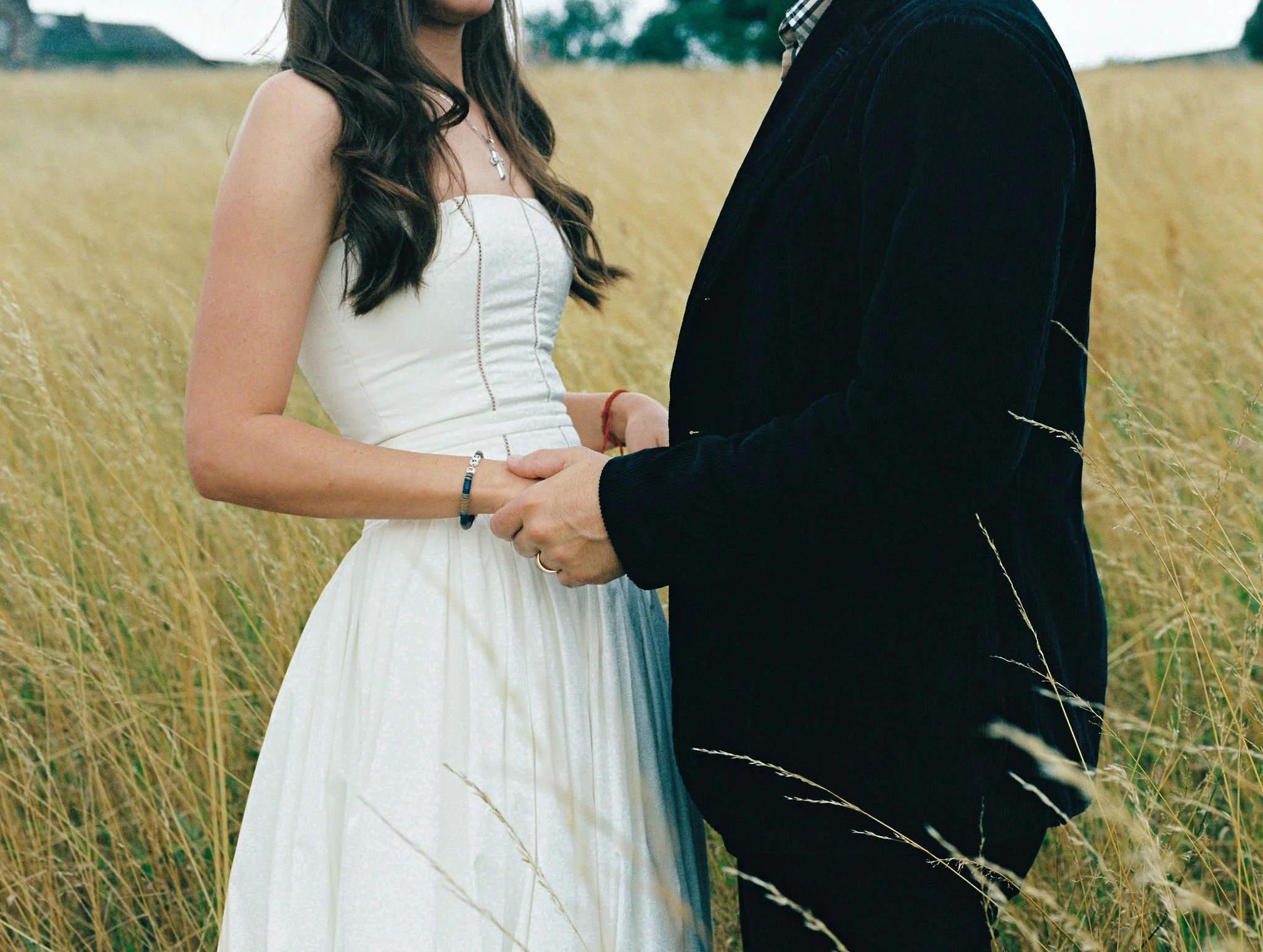 A woman in a white dress holding hands with a man in a dark jacket outdoors in a field of tall grass.