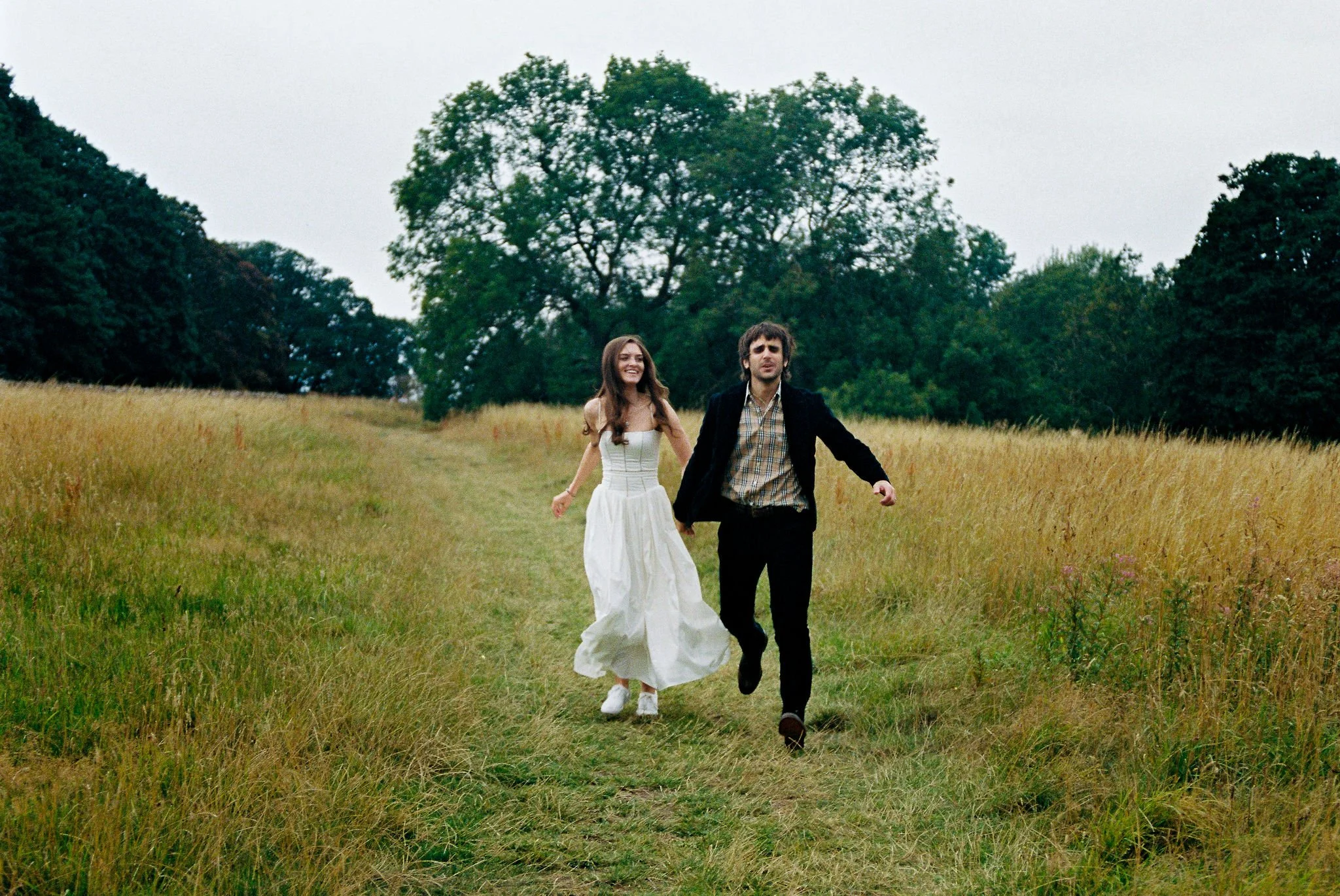 A man and woman running and holding hands in a grassy field with trees in the background. The woman is wearing a white dress, and the man is wearing a dark jacket and plaid shirt.
