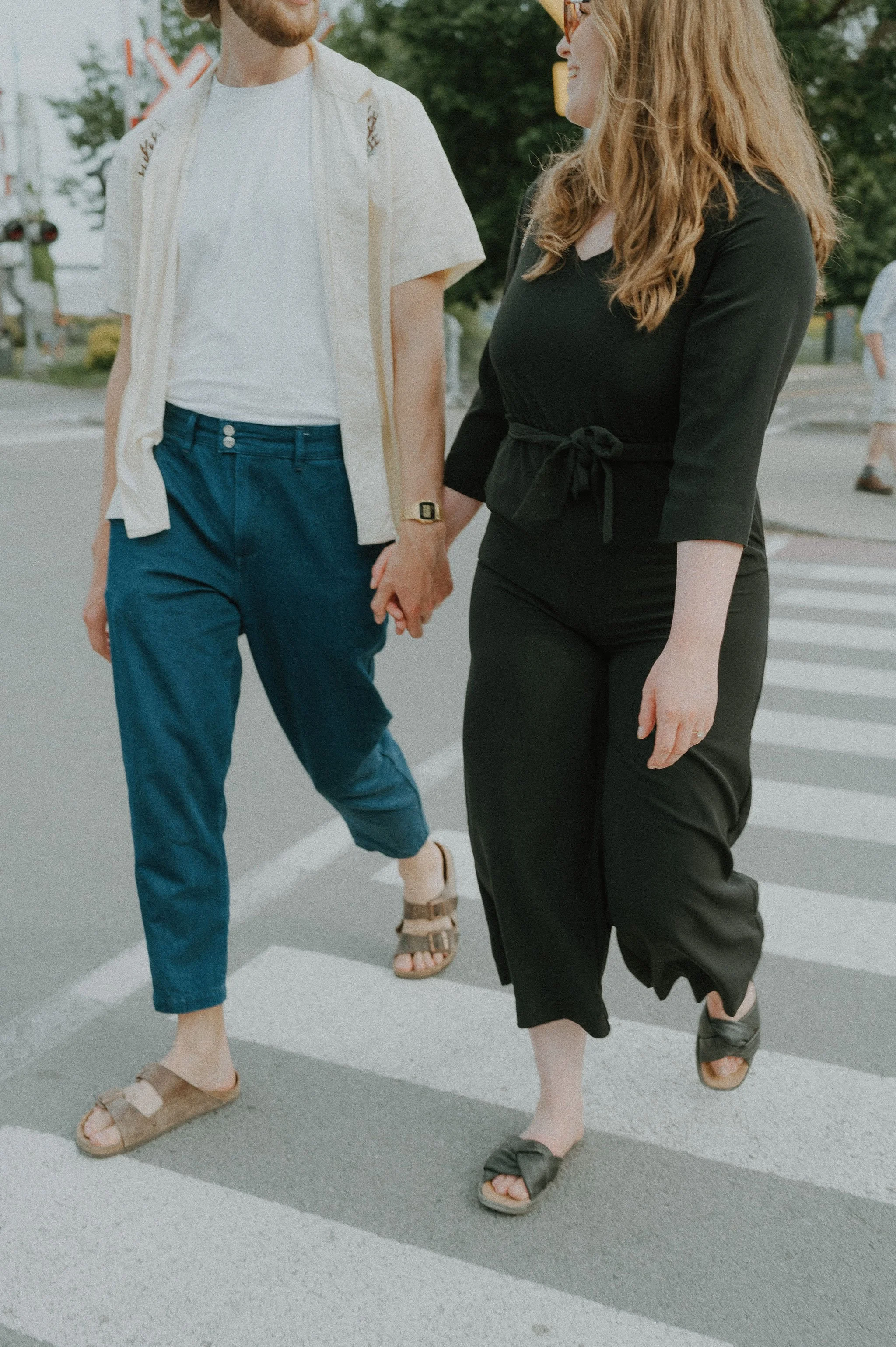 Two people holding hands and crossing a street at a crosswalk, with other pedestrians and trees in the background.