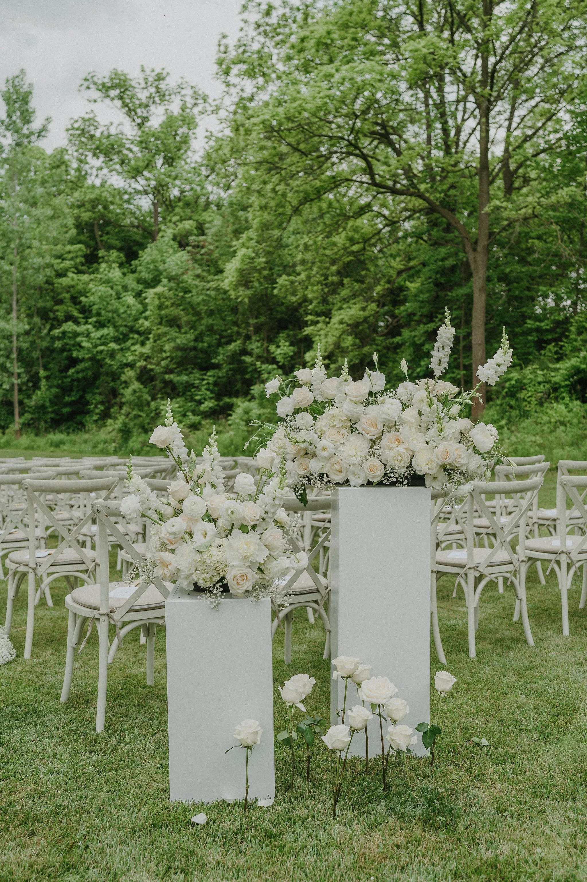 Outdoor wedding ceremony setup with white chairs and large white floral arrangements on white pedestals, surrounded by green trees and grass.