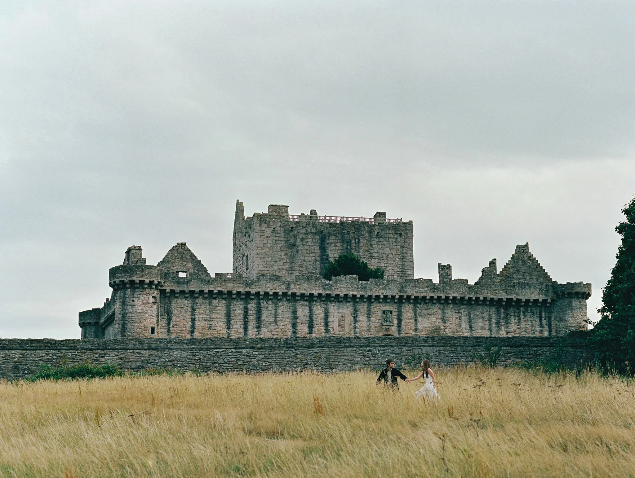 A couple holding hands and dancing in a grassy field in front of an old stone castle with tall walls and towers under a cloudy sky.