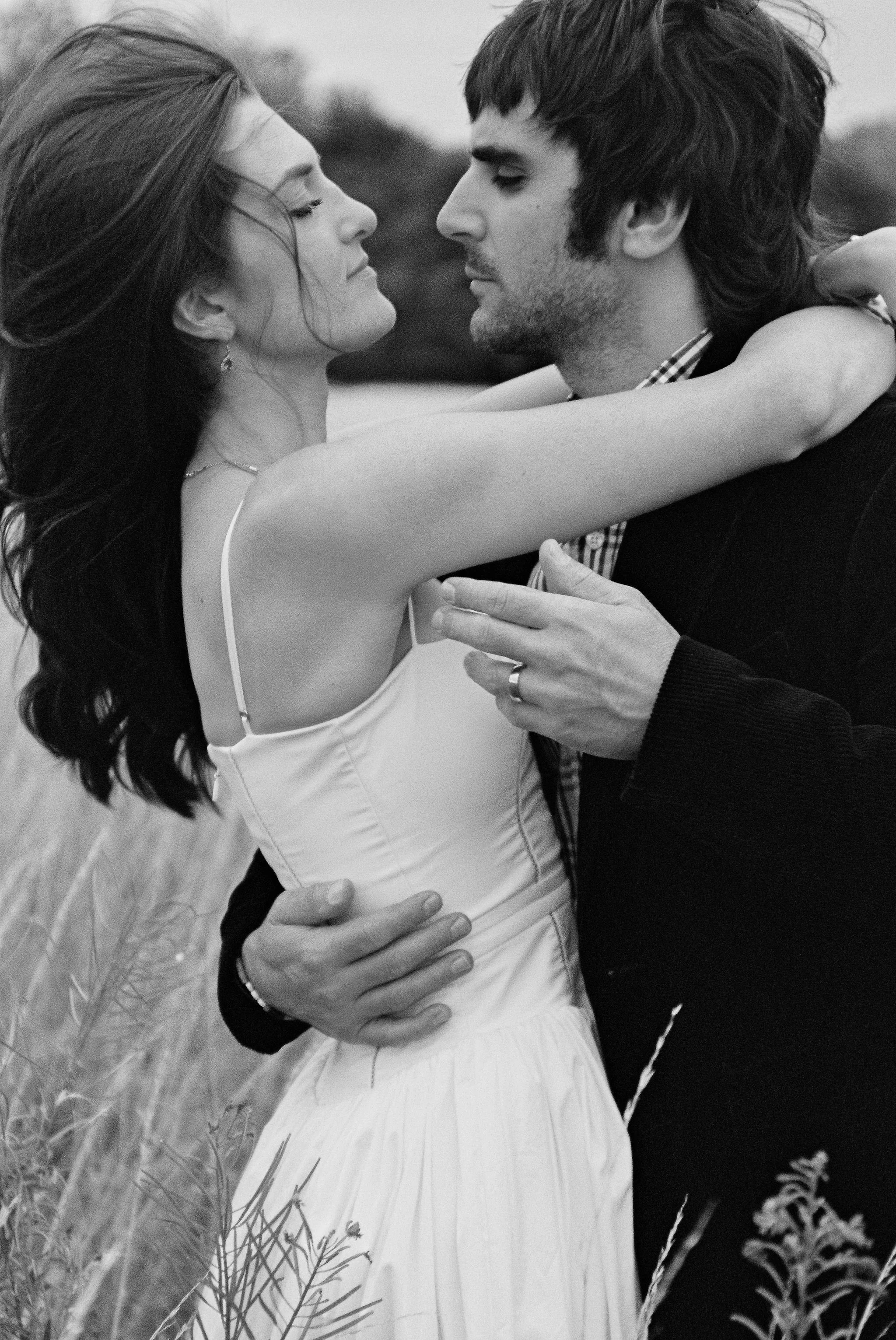 A black-and-white photograph of a couple embracing outdoors, with the woman wearing a spaghetti strap dress and the man in a dark jacket and checkered shirt, close to each other, holding each other tenderly.